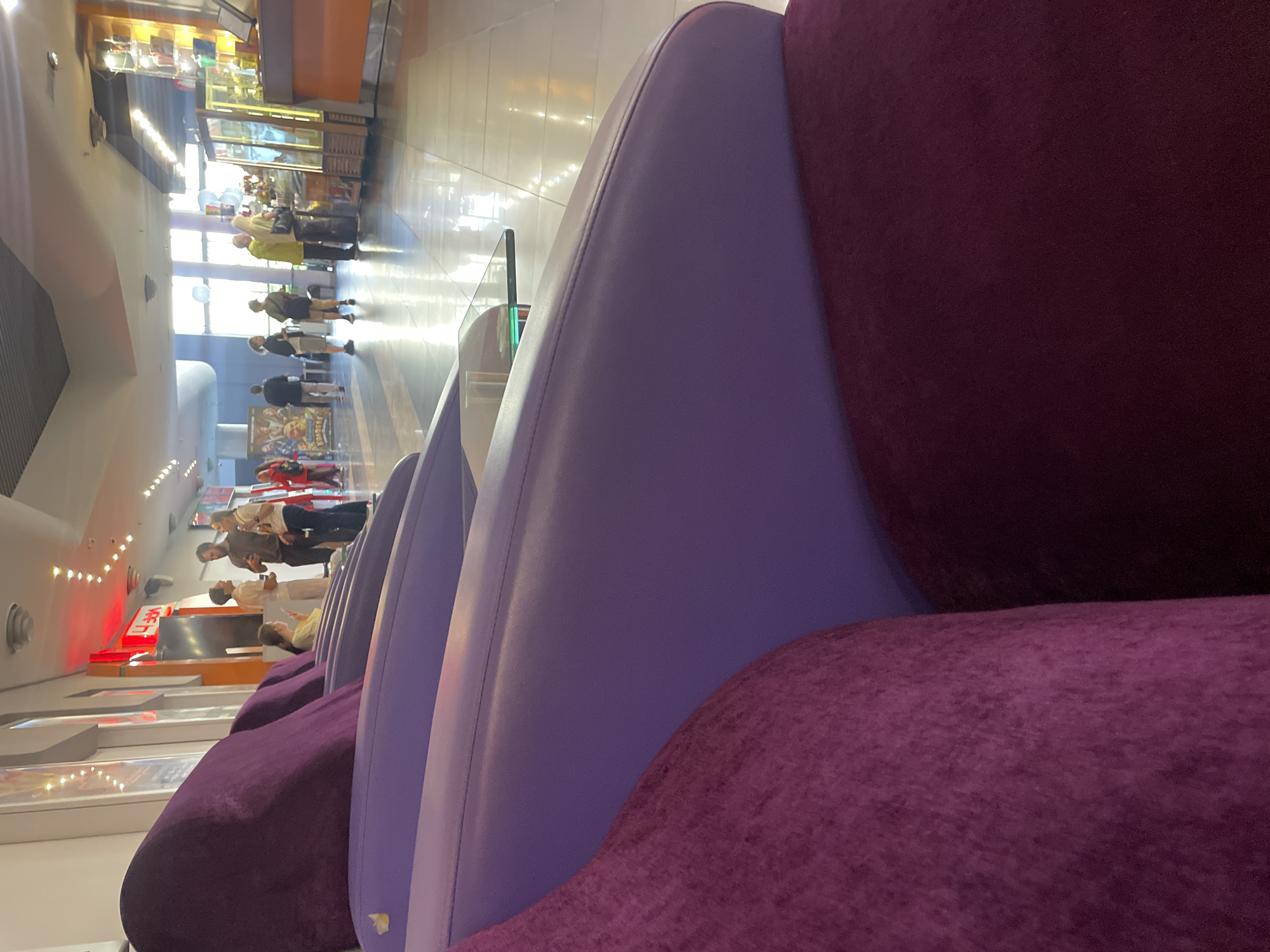 The image shows a dimly lit interior of a cinema lobby. In the foreground, a plush purple sofa is prominently featured, with its textured material and curved design visible. Beyond the sofa, the lobby stretches into the background, featuring a polished tiled floor reflecting the ambient light. Several people are present in the middle ground, appearing to be patrons of the cinema. Some are standing and conversing, while others are walking or looking at displays. Towards the right side of the image, a concessions counter with illuminated display cases is visible, stocked with various items. The overall atmosphere suggests a public space within a cinema complex. The location is Vaganovo, Russia.