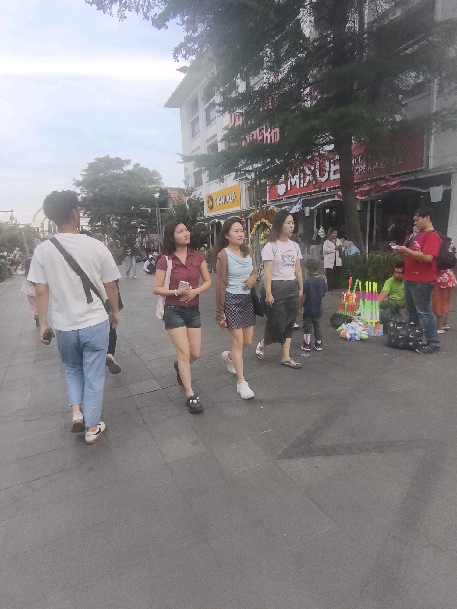 This image captures a street scene, likely in the city of West Jakarta, Indonesia, based on the signage visible. In the foreground, a man wearing a white t-shirt and light blue jeans walks away from the camera. To his right, three women are walking towards the camera. The woman closest to the man wears a red short-sleeved shirt and dark shorts, carrying a light-colored bag over her shoulder. The second woman from the right wears a light blue top and a checkered skirt, while the third woman wears a pink t-shirt and dark pants.
In the background, a storefront with prominent red signage reads "MIXUE," indicating an ice cream and tea shop. Further to the left, another sign reads "TIANLALA." The street appears to be paved with large square tiles. Several other people are visible in the midground and background, some walking and others appearing to be stationary. A street vendor is present on the right side, with a display of colorful items, possibly toys or ice cream-related products. The environment suggests an outdoor urban setting with trees and a multi-story building lining the street. The lighting indicates it is daytime, possibly late afternoon.