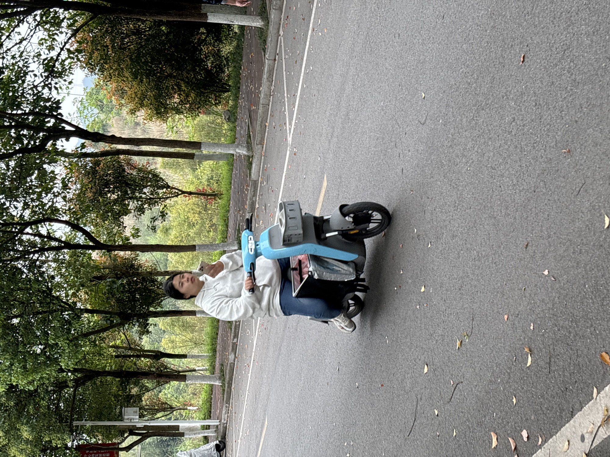 An individual is operating a light blue electric scooter on an asphalt road in Jiangjin, China. The person, dressed in a white hooded sweatshirt, blue jeans, and light-colored sneakers, is positioned centrally on the scooter, holding a light-colored mobile phone in their raised right hand. The electric scooter features a grey front basket and a black rear storage compartment containing red and silver bags. The asphalt road surface exhibits scattered brown and red fallen leaves, along with faint white dashed lines and a yellow line on the left. To the right of the road, a raised concrete sidewalk is visible, also covered with scattered leaves. A row of trees with green foliage and white-painted trunks borders the sidewalk. Beyond these trees, dense green and reddish-brown vegetation covers a sloping background. A white utility pole with a red sign is partially visible on the far left of the sidewalk. The upper arm of another individual is partially visible on the far right of the frame.