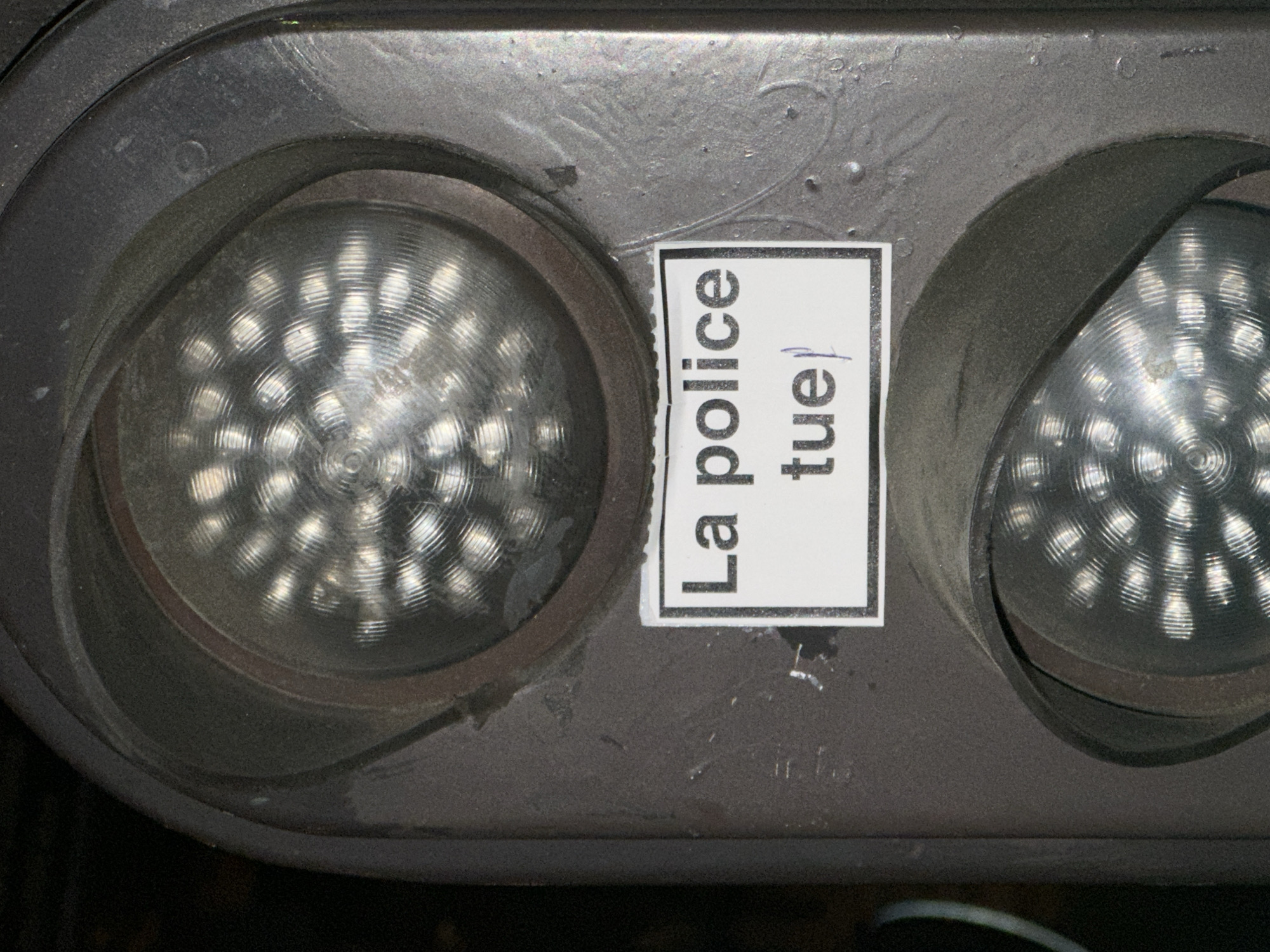 The image displays a close-up of a dark grey traffic signal or pedestrian crossing light, likely located in Paris, France. The signal housing features two circular light fixtures, each containing a grid of multiple small, reflective LED-like elements arranged in a circular pattern, giving them a textured, almost spiraled appearance.
A white rectangular sticker is prominently affixed vertically between the two light fixtures. The sticker has a black border and features black, uppercase French text: "La police tue". Below the word "tue", a small, possibly handwritten, vertical line or character is visible. The phrase translates to "The police kill."
The traffic signal's surface appears weathered, with scuffs and minor abrasions visible on the dark grey casing, particularly around the edges of the light compartments. The lighting suggests a low-light environment, possibly evening or night, enhancing the reflectivity of the light elements. There is no human activity or specific event depicted, focusing solely on the urban fixture and its added message.