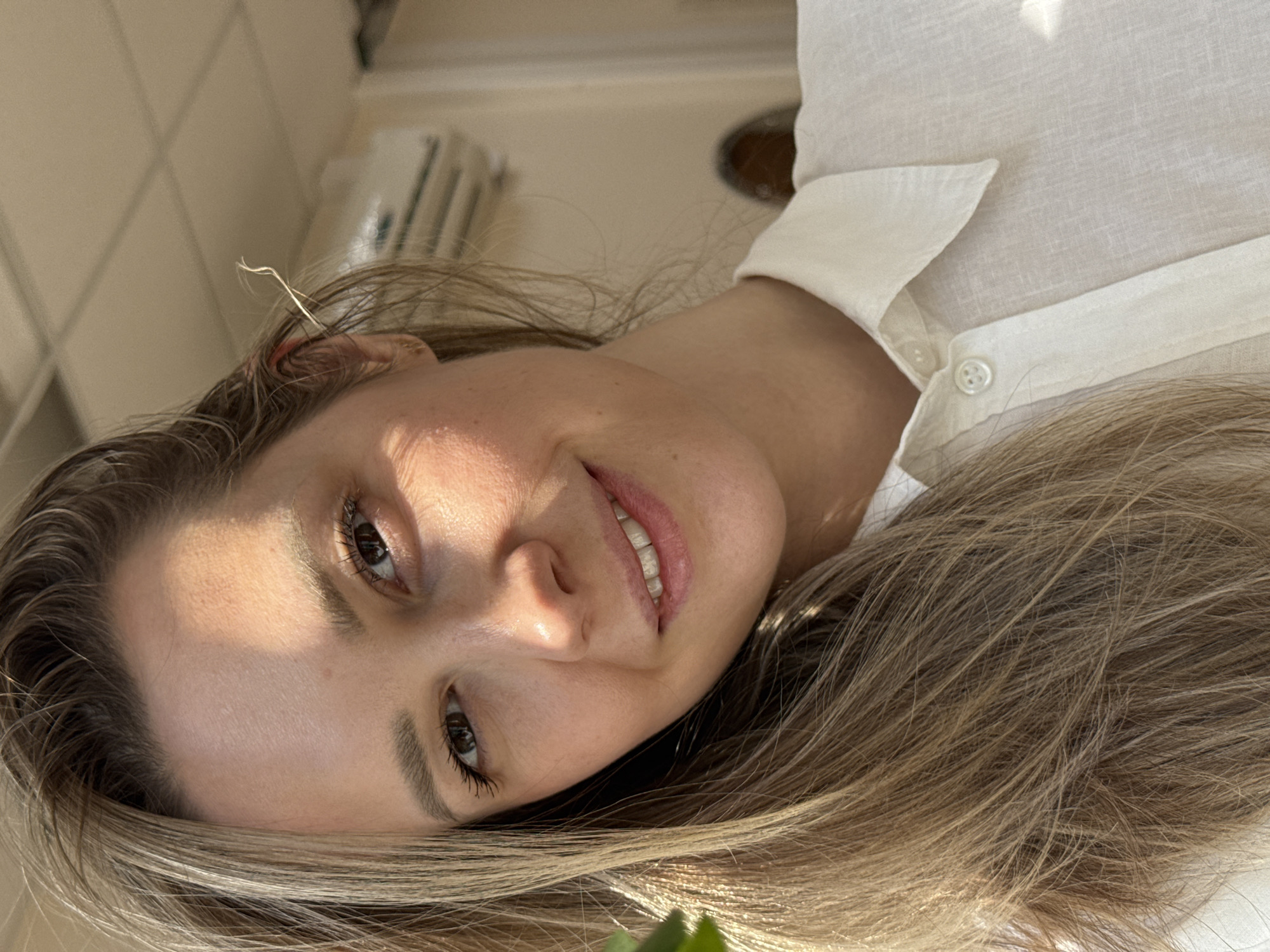 A young woman with long, highlighted brown hair smiles slightly at the camera. She is wearing a white linen shirt. The photo appears to be taken indoors in a brightly lit room, with natural light illuminating her face. The background is blurred, but a portion of a white appliance or fixture is visible behind her. The location in the city of Chaikovsky, Russia is not discernible from the image.