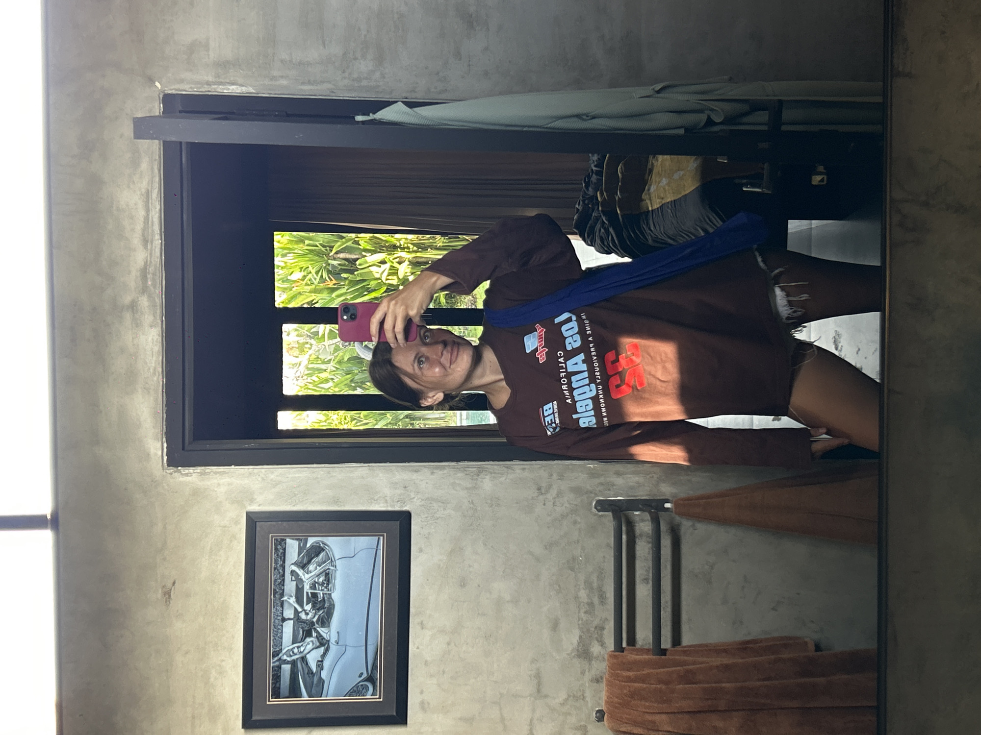 A woman is reflected in a mirror indoors. She is holding a pink phone, presumably taking the photo. The woman is wearing a long-sleeve brown shirt with red lettering and dark blue garment around her neck. The room is concrete with a black-framed painting of a car and a towel rack. There is greenery visible through a window in the background. The location is Mengwi, Indonesia.