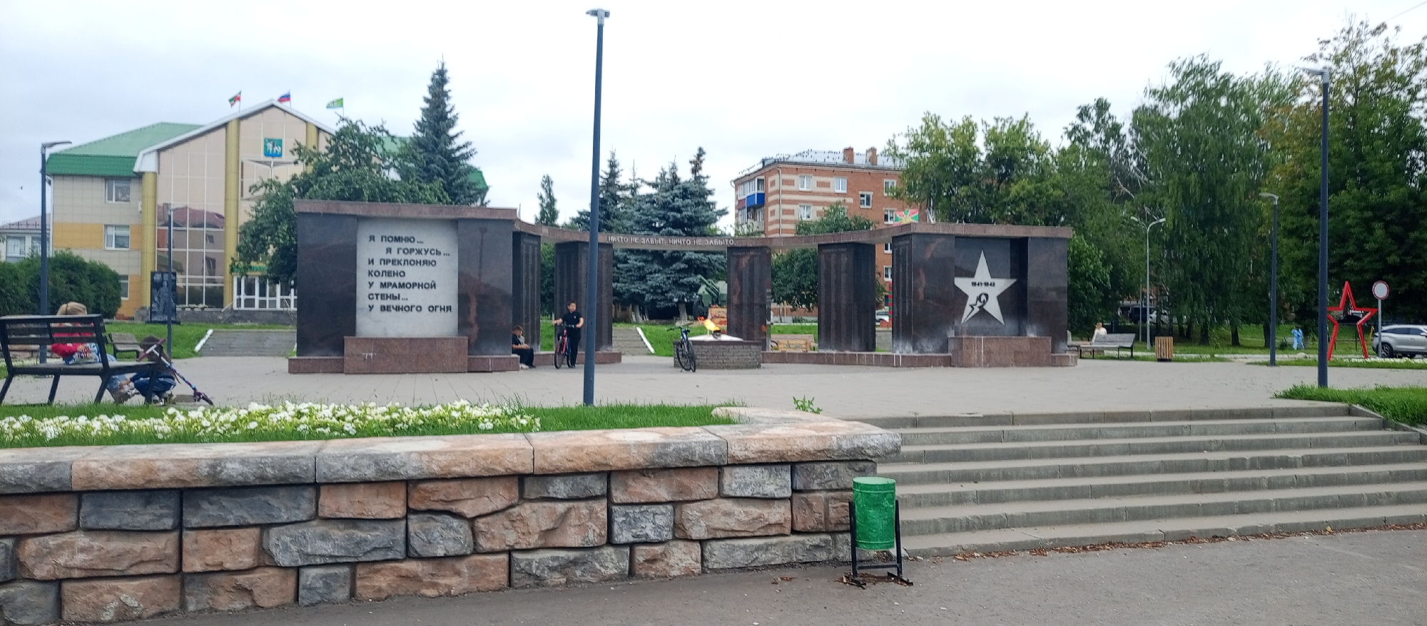 The image depicts a public square in Agryz, Russia, featuring a memorial complex. The memorial is constructed of dark stone and includes several upright panels inscribed with text and symbols. Prominently displayed on one panel is a large white star with the years "1941-1945" within it, indicating a war memorial. A permanent flame burns at the base of another section of the memorial, where two bicycles are parked.
In the foreground, a low wall made of multi-colored stones separates the paved area from a grassy strip with white flowers. To the left of the memorial, a young child on a scooter is playing near a person sitting on a park bench. A light-colored building with a green roof is visible in the background, along with several tall evergreen trees and other deciduous trees. The sky is overcast. Further to the right, a green trash can stands near a set of concrete stairs leading up to the square. A red star sculpture is also visible in the background to the right of the memorial.