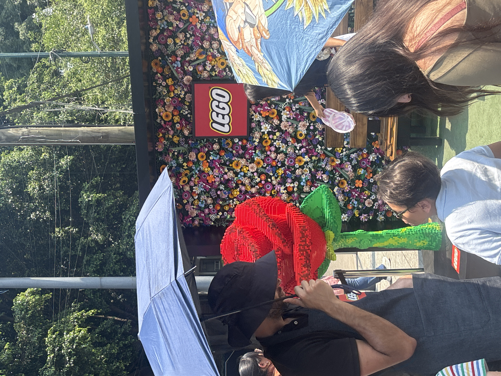 The image shows a LEGO installation in what appears to be Mexico City, Mexico. A large LEGO rose is in the foreground, with people looking at it. Behind the rose, there is a wall of flowers with a LEGO logo. People are holding umbrellas, suggesting it might be raining or sunny. The environment includes trees and potentially a park.