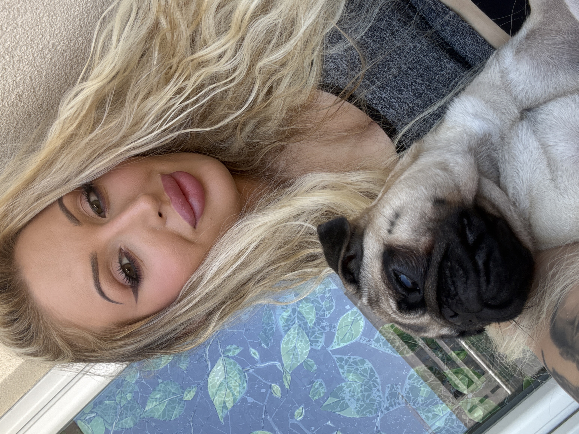 A close-up selfie shows a blonde woman with full lips and heavy eye makeup holding a fawn-colored pug. The woman's head is to the right of the frame, and she is wearing a gray top. The pug is in the lower left quadrant of the image, nestled in the woman's arms. The background consists of a blue wall with a green leaf pattern, possibly a window covering or mural. The lighting suggests an outdoor setting.