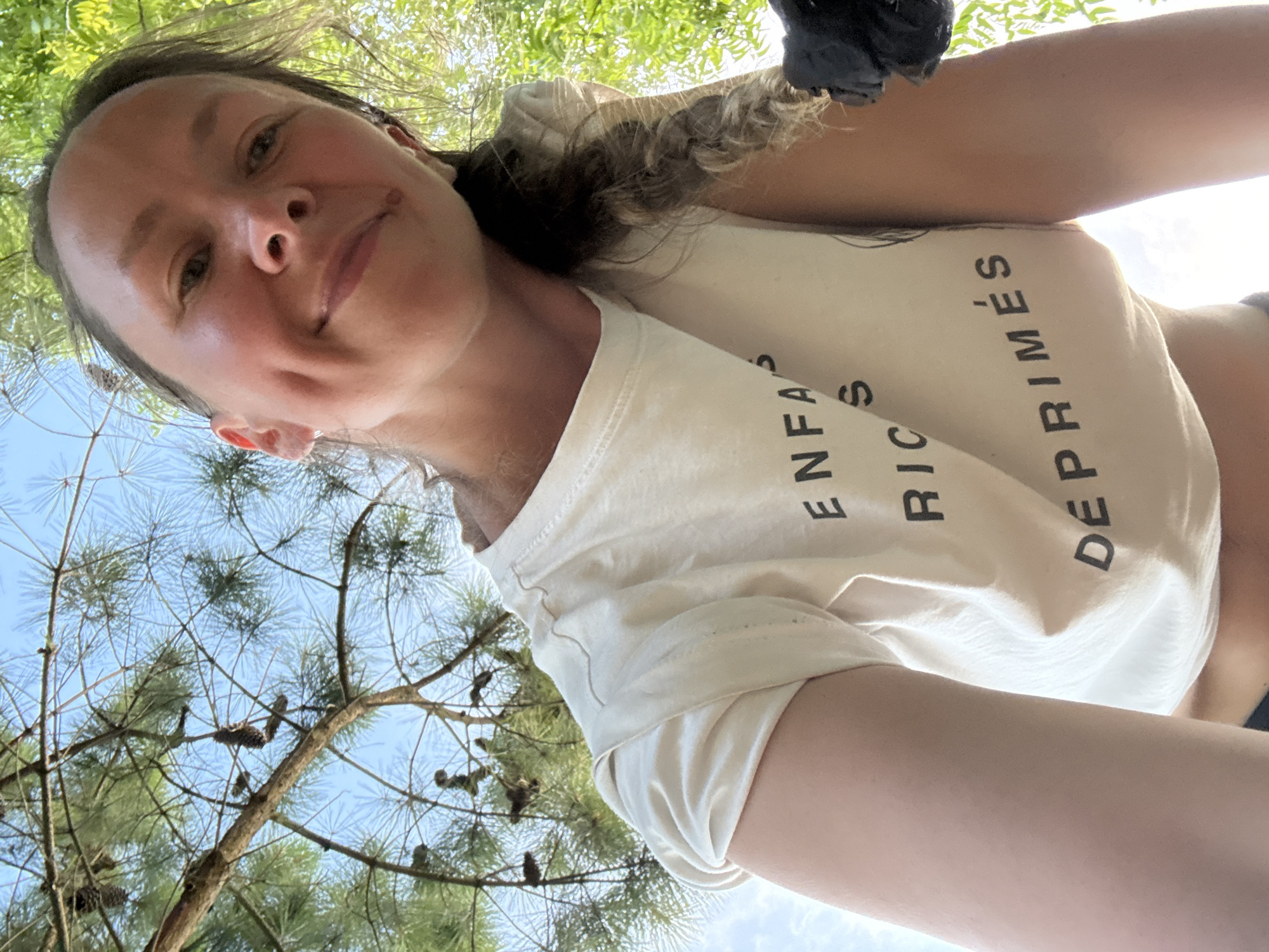A woman is captured in a low-angle, outdoor selfie. She is wearing a cream-colored cropped t-shirt with black text printed on it that reads "ENFANTS DEPRIME(S)". Her left arm is raised, and the image is taken from a perspective looking up at her. The background consists of lush green foliage and a bright blue sky, suggesting it is daytime and the weather is clear. Pine needles and pinecones are visible in the branches of a tree in the foreground, hinting at a natural, possibly forested environment. The woman has a friendly smile and her hair is pulled back in a braid.