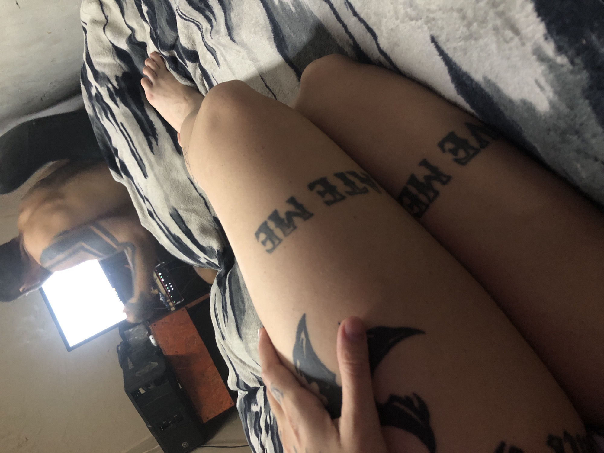 The image shows a person lying on a bed with patterned bedding. Tattoos are visible on their thighs, with the words "HATE ME" on one thigh and "LOVE ME" on the other. A second person is partially visible in the background, shirtless and leaning over what appears to be a computer setup. The setting appears to be indoors, possibly a bedroom, with a computer tower and screen visible. It is not possible to determine if the image was taken in Buenos Aires, Argentina.