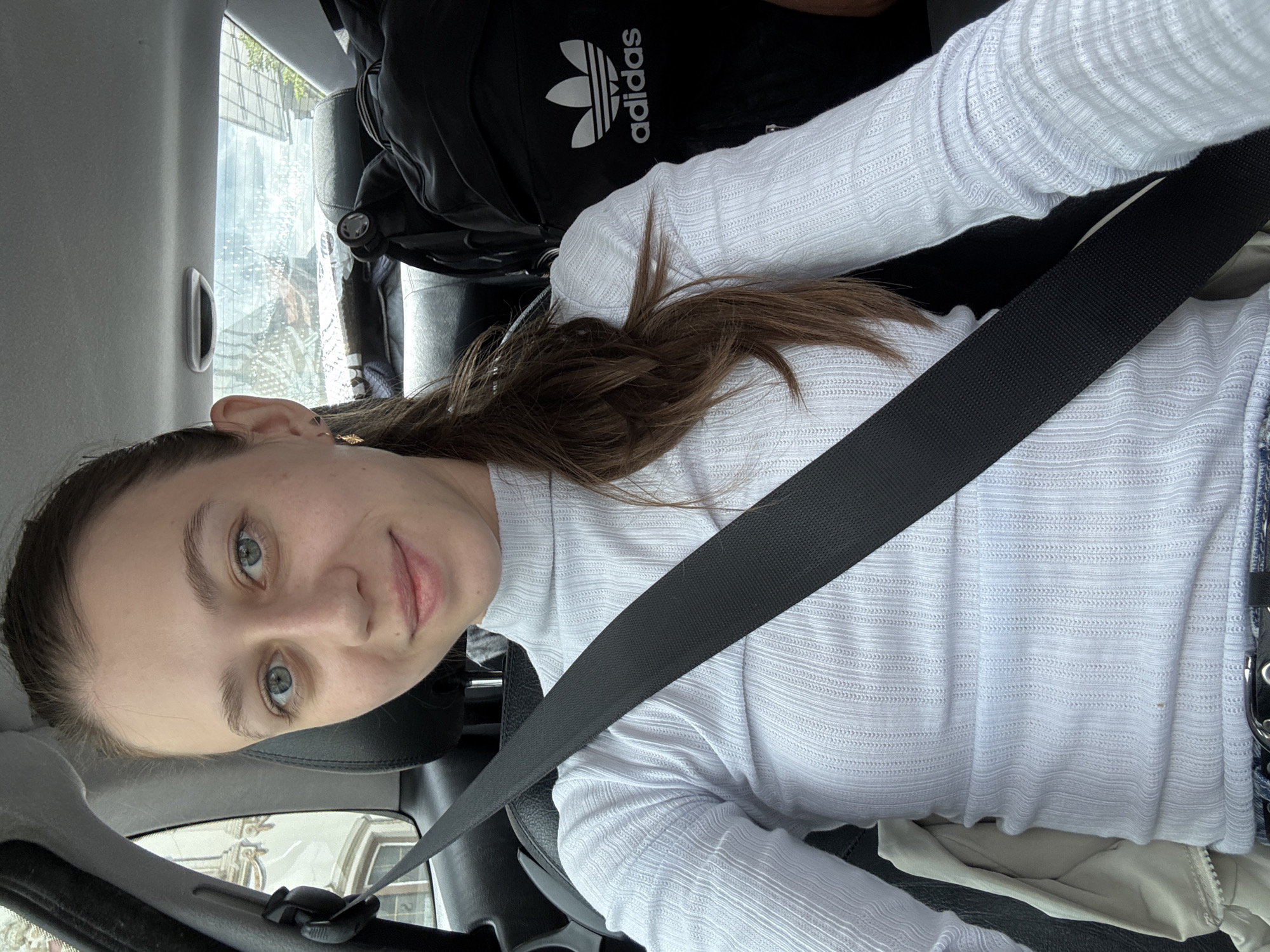 In the city of Reutte, Austria, a young Caucasian woman with light brown hair, blue eyes, and fair skin is photographed from the chest up inside a car. She is wearing a white, long-sleeved, ribbed turtleneck sweater and a black seatbelt is fastened across her chest. She is looking directly at the camera with a slight smile. In the background, a black backpack with the Adidas logo is visible on the car seat behind her. The car's interior, including the headrest and ceiling, is also visible.