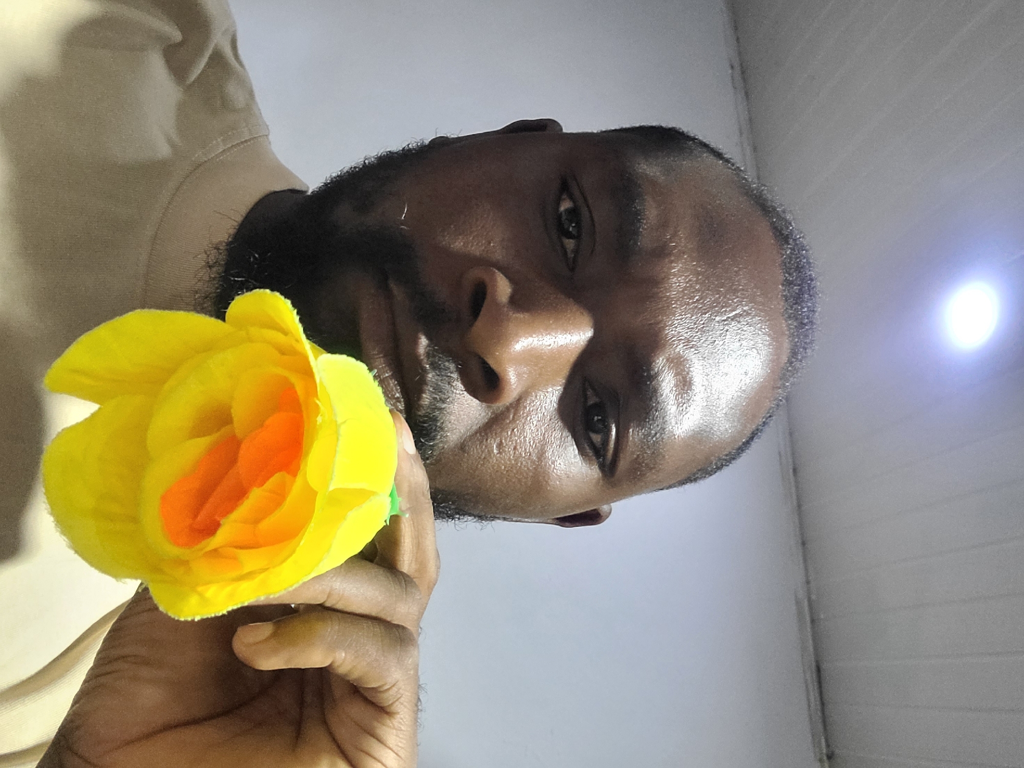 A medium, angled, close-up shot shows a dark-skinned man with a short black beard holding a yellow and orange rose. The man is wearing a beige t-shirt. The background is a light grey wall. No specific details indicating the location as Jos, Nigeria are visible.
