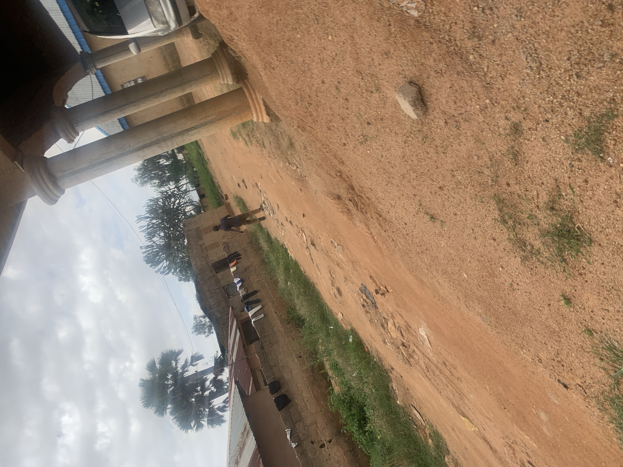 The image depicts a rural or semi-urban scene in Du, Nigeria, characterized by a dirt road and traditional brick structures. In the foreground, a sandy, orange-brown dirt path slopes upwards. To the left, a low-lying wall constructed from mud bricks separates the road from a property. Clothes are hanging on a line strung along the top of this wall, including shirts, pants, and what appear to be socks or underwear. Beyond the wall, a building with a corrugated metal roof is visible.
Further back, a person, appearing to be a male, is walking away from the camera on the dirt road. He is wearing a dark shirt and light brown or khaki pants. The background reveals more of the brick buildings, some with what appear to be simple windows. Trees, including palm trees and some spiky plants, are scattered throughout the scene, suggesting a warm climate. The sky is overcast with grey clouds. To the right of the scene, large, decorative concrete pillars suggest a more substantial structure, possibly a veranda or entrance to a building, with a silver vehicle partially visible behind them. The overall impression is one of a modest, lived-in environment.