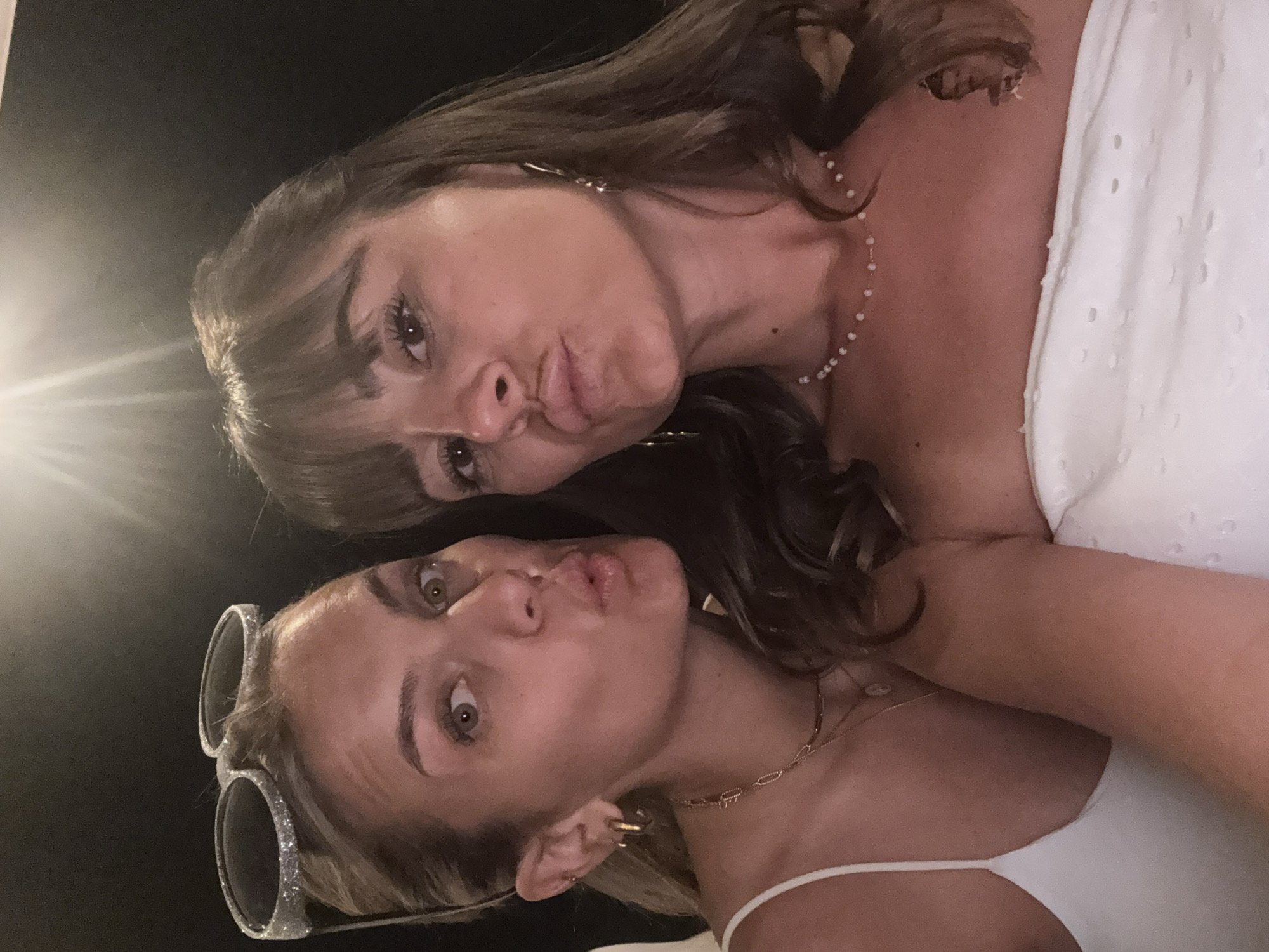 Two women are taking a selfie. The woman on the left has blonde hair pulled back and is wearing a white tank top and a necklace. She has sunglasses on top of her head. The woman on the right has brown hair and is wearing a strapless white top and a beaded necklace. Both women are puckering their lips. The background is dark, and there is a bright light source from above and to the left. The location is specified as Savigneux, France.