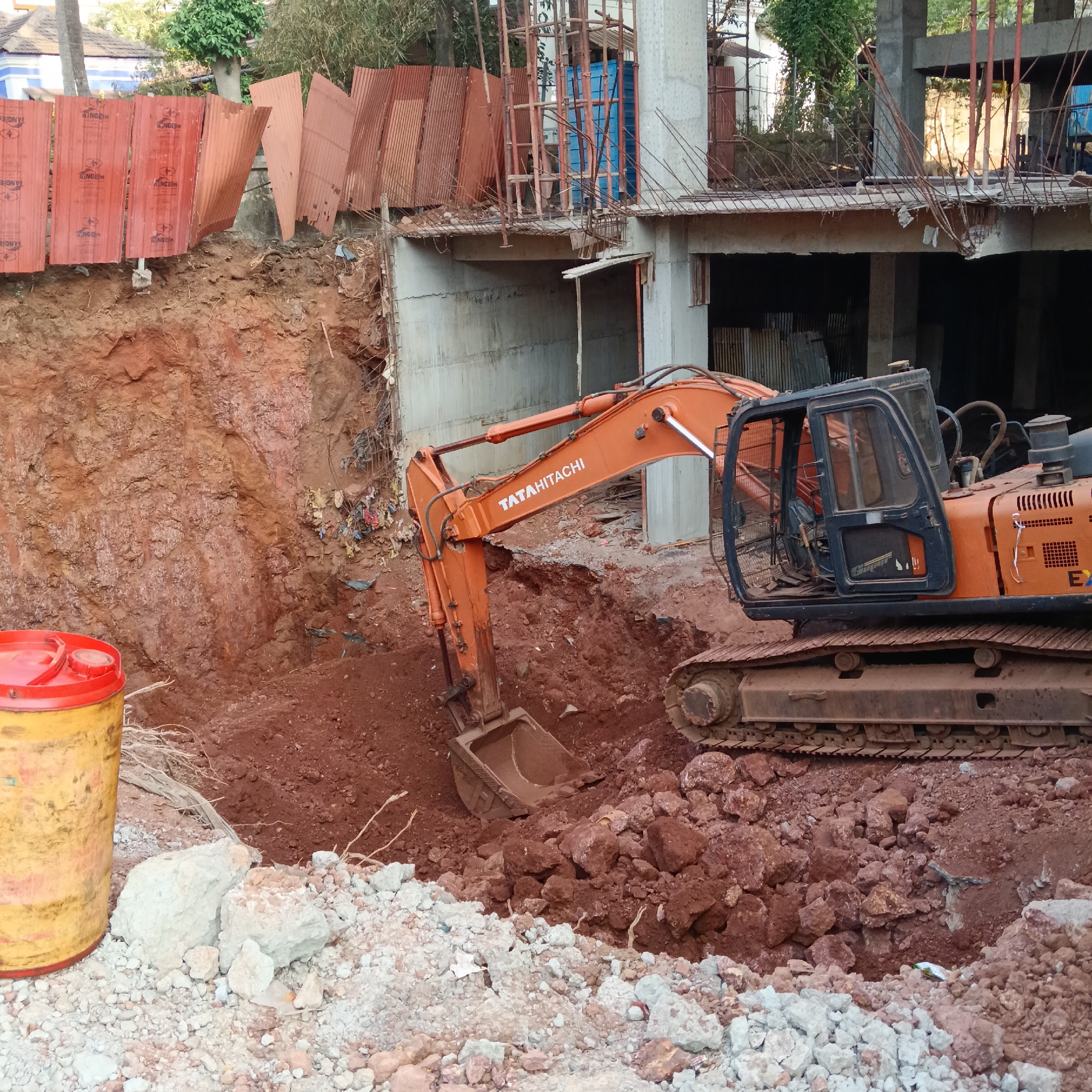 The image depicts a construction site in what appears to be a tropical or subtropical region. The scene is dominated by a large orange Tata Hitachi excavator actively digging into a pit of reddish-brown soil. A barrel, yellow with a red lid, sits to the left near the edge of the pit, among a pile of light-colored rocks and debris. Behind the pit is a raw earth wall showing signs of digging and excavation.
In the background, a partially constructed concrete building is visible. Scaffolding and exposed rebar are present. Red corrugated metal sheets lean against the edge of the pit. The overall impression is of ongoing excavation work for building construction. The location information (Mapusa, India) provides further context, indicating a construction project in a specific geographical area. No people are clearly visible in the scene. The primary subjects are the construction equipment, building under construction, and excavated earth. The action is the excavator digging into the soil.