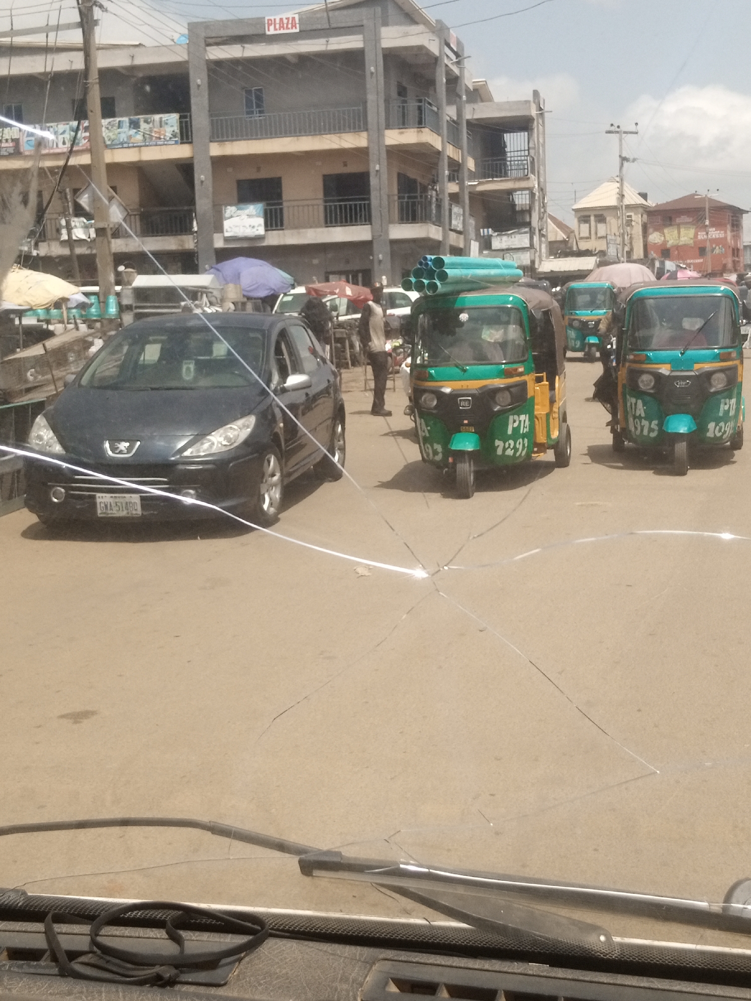 This media file captures a bustling street scene in Jos, Nigeria, under a bright, likely midday sky with some clouds. The foreground shows the cracked windshield of a vehicle, with a wiper blade visible. Beyond the windshield, the street is filled with various vehicles. A dark grey Peugeot sedan with license plate "GWA 51480" is prominently featured on the left. To its right are several green and yellow auto-rickshaws, commonly known as "Keke Napep" or "Marwa" in Nigeria.
One of these auto-rickshaws is carrying a stack of light blue PVC pipes on its roof. Another has "PTA 7293" visible on its side, while a third displays "PTA 1975" and "PTA 109". The auto-rickshaws appear to be in motion or waiting, with drivers and possibly passengers inside. In the background, multi-story buildings line the street, some with signs. One building has "PLAZA" written in red letters. The overall atmosphere suggests a typical urban marketplace or transit hub, characterized by activity and the presence of various modes of transportation. The presence of the auto-rickshaws and the style of the buildings are strong indicators of a Nigerian location.