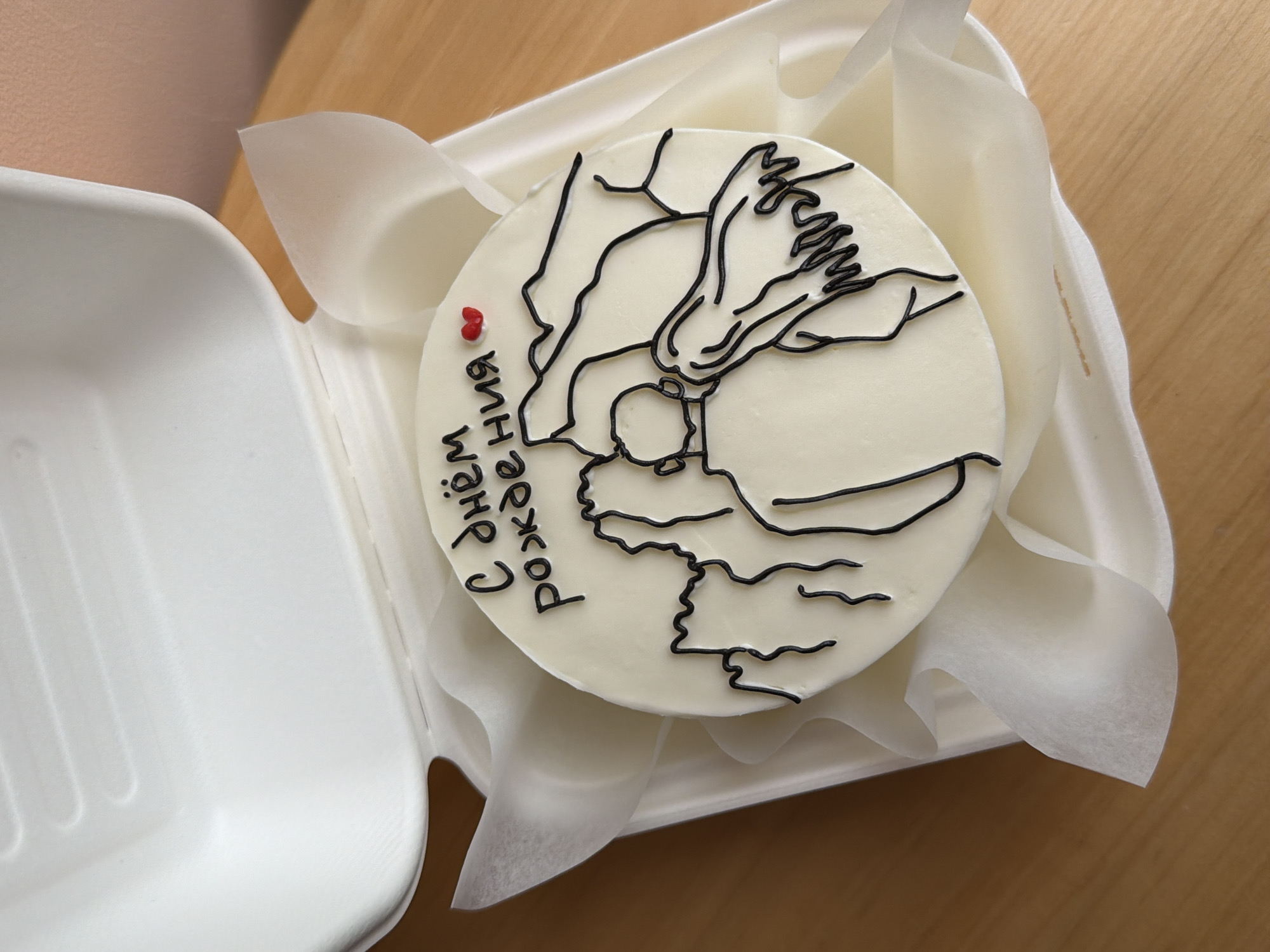 A top-down shot shows a small, round cake decorated with a black line drawing of what appears to be a person embracing a child, and the Russian phrase "С днем рождения" ("Happy Birthday"), with a small red heart. The cake is resting in a white, rectangular container, cushioned by white tissue paper. The container sits on a light brown, wooden surface. The location of Tel-Aviv, Israel, cannot be determined from the context of this image.