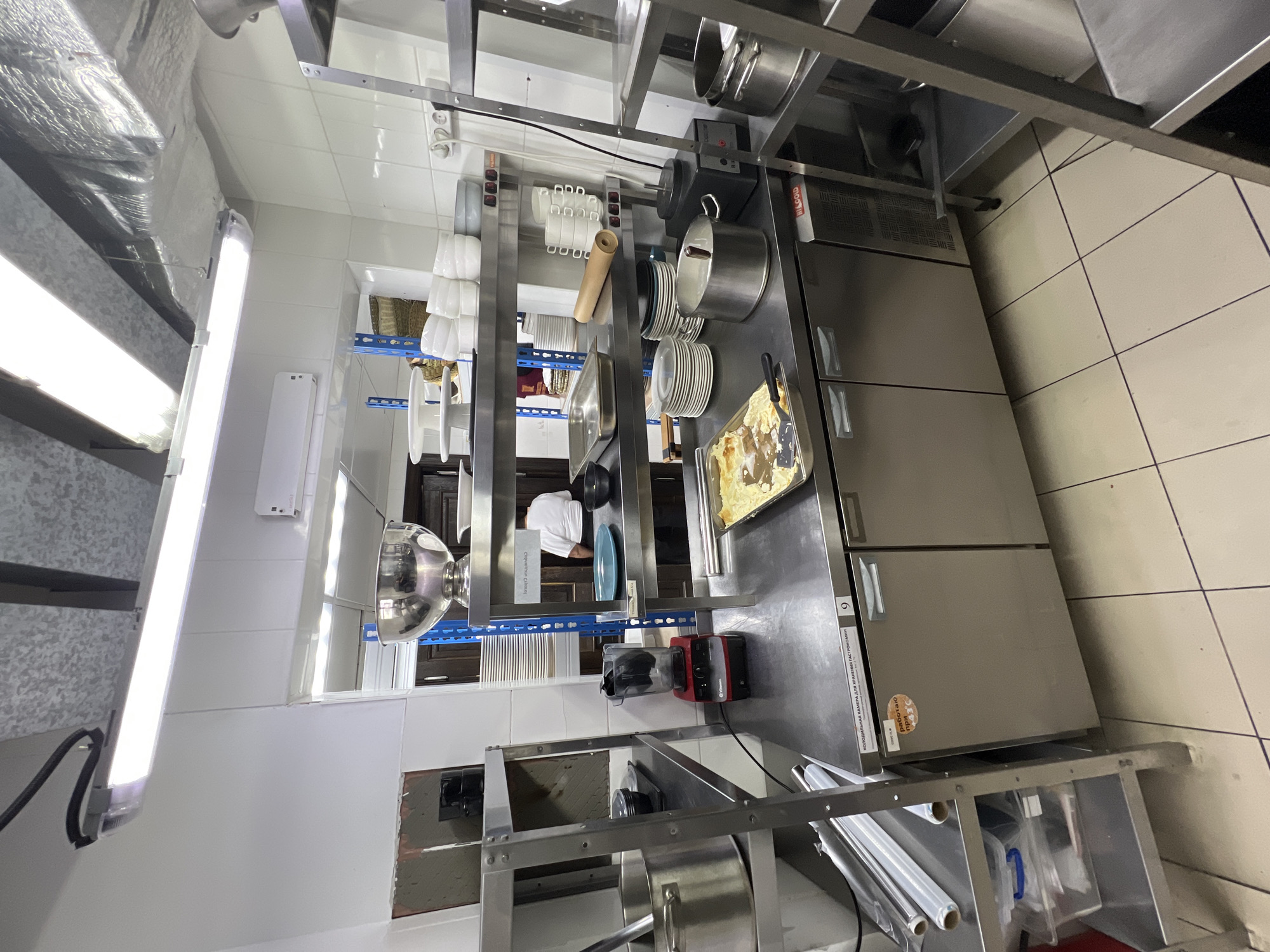 The image depicts a commercial kitchen. In the foreground, a stainless steel countertop contains various kitchen equipment, including a blender, a large pot, and a baking tray with food. To the right, a stainless steel refrigerator unit is visible. Behind the countertop, metal shelving units are stacked with plates, bowls, and other kitchenware. A chef in a white uniform stands partially visible in the background. The overall environment is clean and organized, suggesting an active food preparation area. The image does not provide any information about the city of Plast univska, Russia.