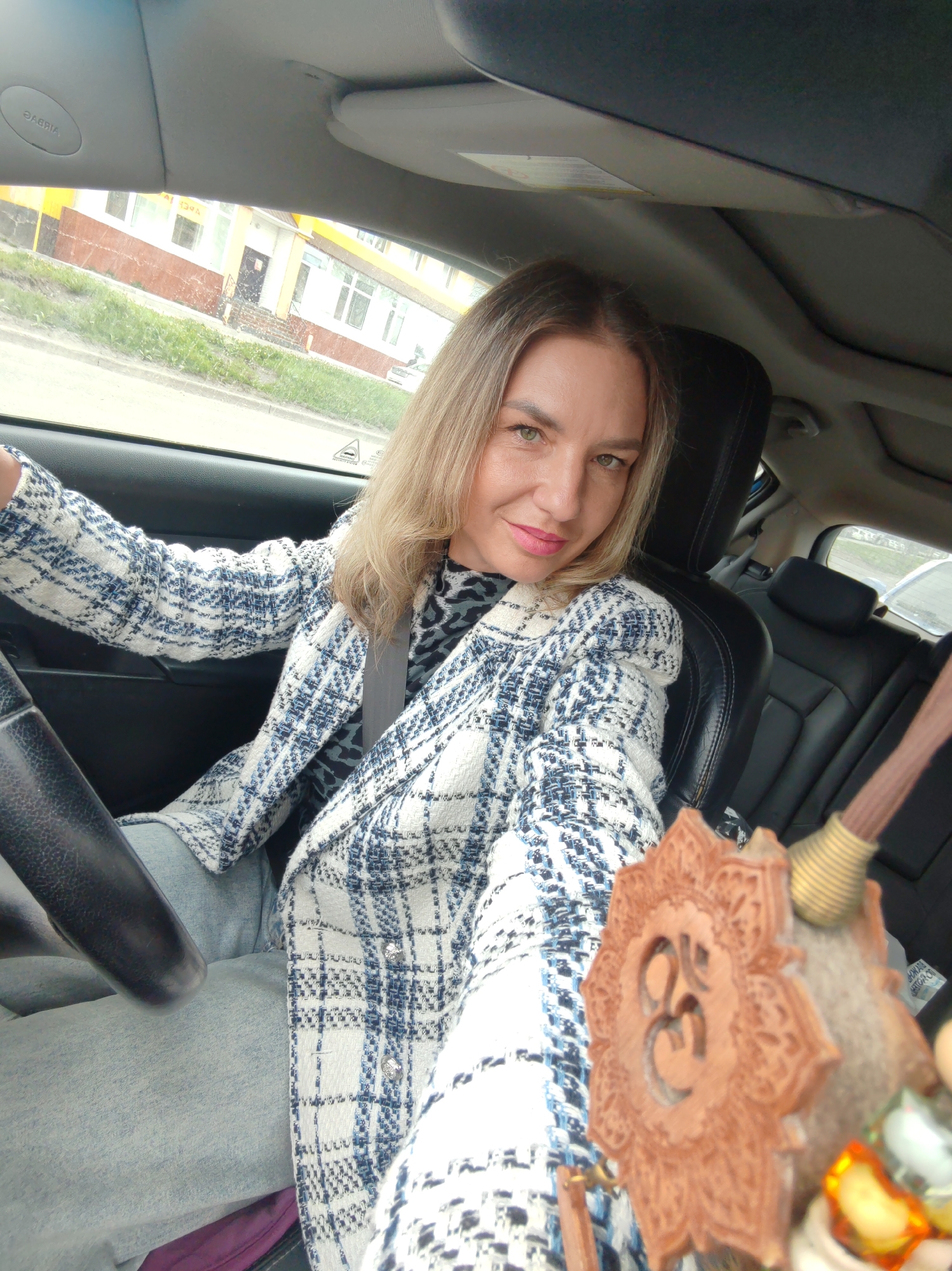 A blonde woman with green eyes and pink lipstick is sitting in the driver's seat of a car. She is wearing a blue and white checkered jacket over a leopard-print top and light-colored jeans. Her right hand is on the steering wheel, and her left arm is extended towards the camera. In the foreground, a wooden amulet with an "Om" symbol is visible, along with what appears to be an incense stick holder. Through the car window, a building with a yellow facade and red trim is visible, along with grass and a sidewalk. The car's interior includes black leather seats and a sunroof. The scene is consistent with a street view in Petropavlovsk-Kamchatsky, Russia, where similar architecture and car models are common.