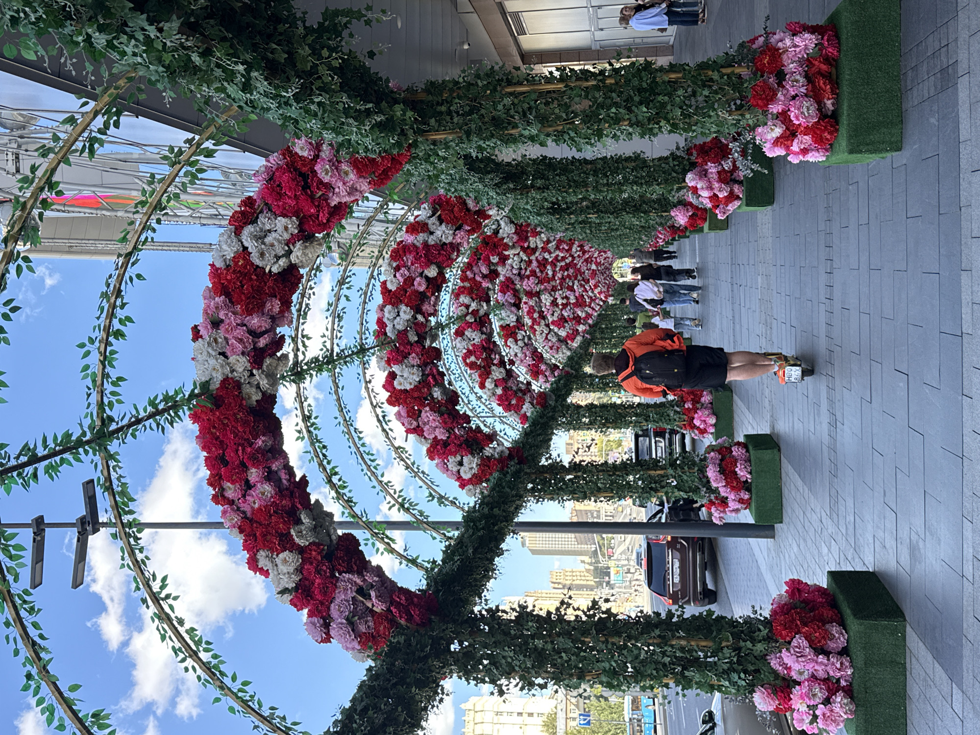 The image shows a pedestrian walkway in Moscow, Russia, featuring a decorative archway constructed of artificial greenery and artificial flowers in shades of red, pink, and white. The walkway is paved with gray tiles, and on the left side, there is a street with traffic, including a dark red SUV and a black car. Buildings line the right side of the walkway. Several people are visible: a person wearing an orange jacket and black shorts is riding an electric scooter away from the camera, heading down the walkway. Further down the walkway, a group of people, including adults and a child, are walking. On the far right, two women are standing. The sky is blue with scattered clouds. The overall scene suggests an urban environment decorated for an event or festive occasion.