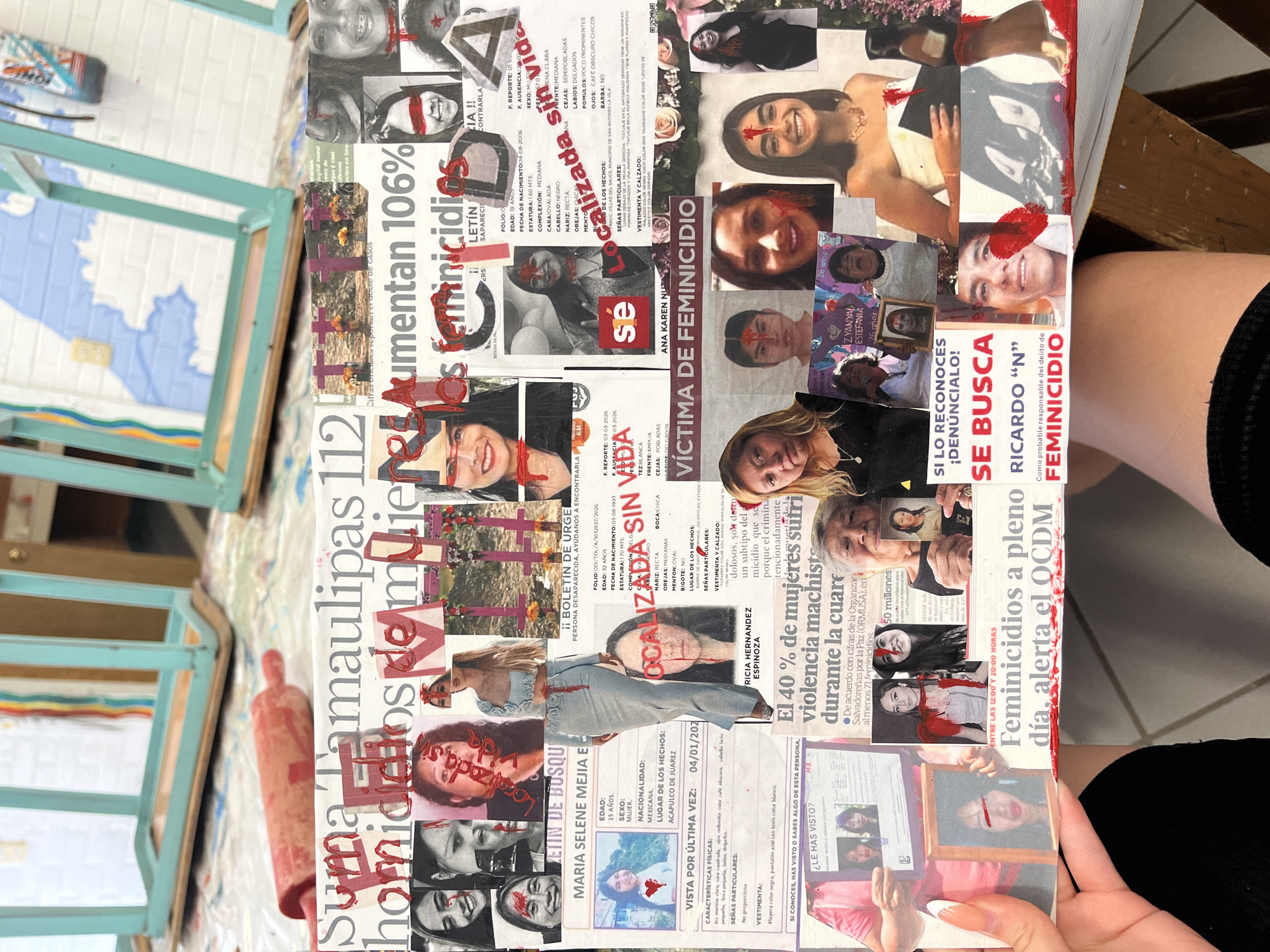 A collage of newspaper clippings and photos is displayed on a surface, possibly a table or canvas, held by a person's hand. The clippings depict images of women and text related to violence against women. The collage includes headlines like "Aumentan 106% femicidios" and "Víctima de Femicidio," suggesting a theme of femicide. Many faces in the collage are marked with a red, blood-like paint. There are also official-looking documents like "boletin de urge" (urgent bulletin) and descriptions of missing women. The environment includes other art supplies, such as a paint roller and painted chairs. The text is in Spanish. There is no indication the scene is located in Puebla, Mexico.
