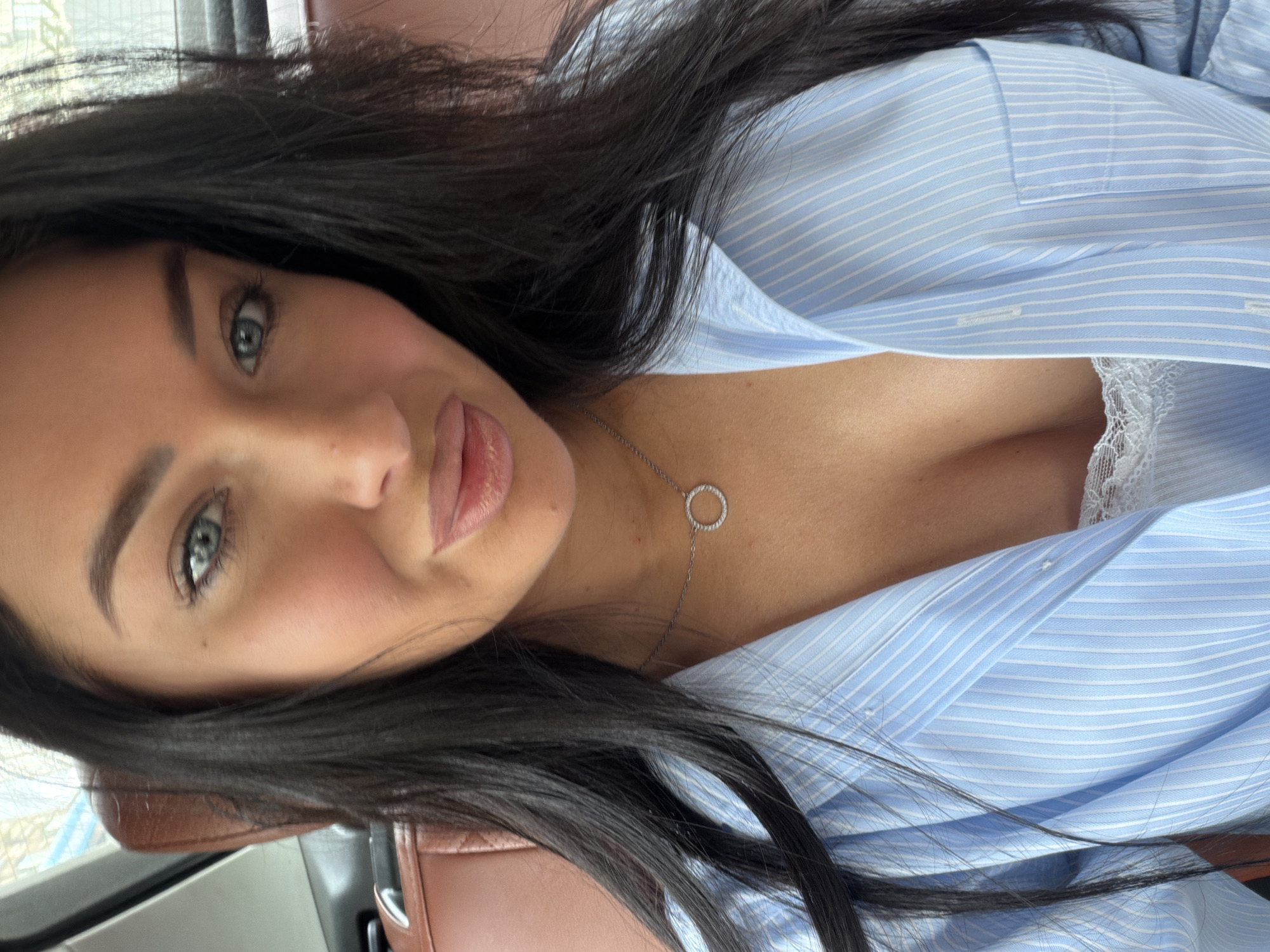 A woman with long, dark hair and blue eyes is looking at the camera. She is wearing a light blue pinstriped button-down shirt, partially unbuttoned, revealing a white lacy camisole underneath. A silver necklace with a circular pendant is visible around her neck. She is seated in what appears to be the back seat of a car, with a brown leather seat visible behind her. The background is blurred, suggesting movement or an out-of-focus environment. There is no indication of the specific location within Moscow, Russia.