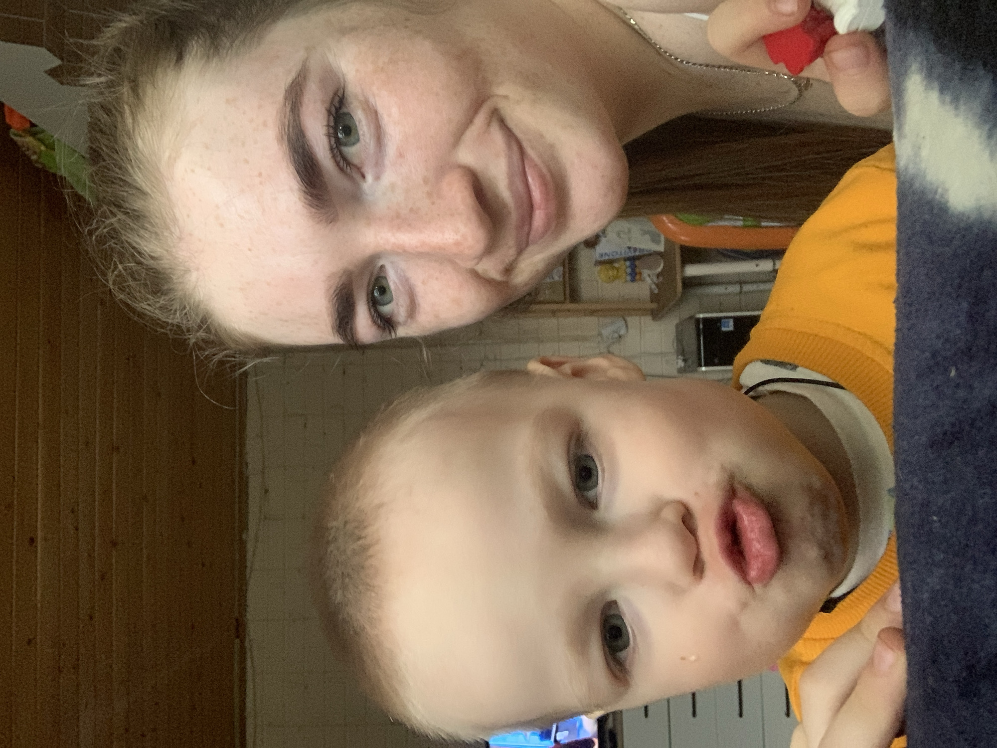 An image, oriented vertically, captures a close-up of a young woman and a child, likely indoors in Chaikovsky, Russia. The woman, positioned at the top left of the frame (when upright), has light hair, freckles, and light green eyes, smiling gently. She wears a light top and a thin chain necklace. Her right hand, visible on the right side of the frame, holds a small red and white object, possibly a berry or a piece of candy.
Below her and slightly to the right is a young child, possibly a boy, with light, short hair and light-colored eyes. His mouth is pursed, and there are distinct dark smudges around his lips and chin, suggesting he has recently eaten something like chocolate or berries. He wears an orange long-sleeved top with a white collar.
The background is an indoor setting. To the left, wooden paneling covers a wall. The right side of the background shows a blurry dark blue and white object, possibly a blanket or furniture. Further in the background, a shelf or cabinet holds various items, with the partial word "STONE" visible on a box. The overall impression is an intimate family moment.