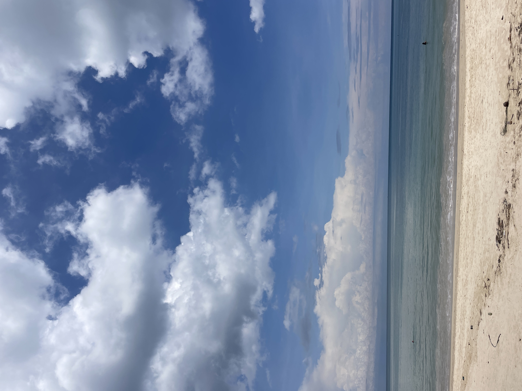 The image depicts a beach scene in Choeng Thale, Thailand. The sky is a mix of deep blue and puffy white clouds. Below the sky is the ocean with calm waves near the sandy beach. A single person is wading in the ocean in the distance. The sand is light tan, with some sparse debris, including a few small branches. The scene is oriented horizontally.