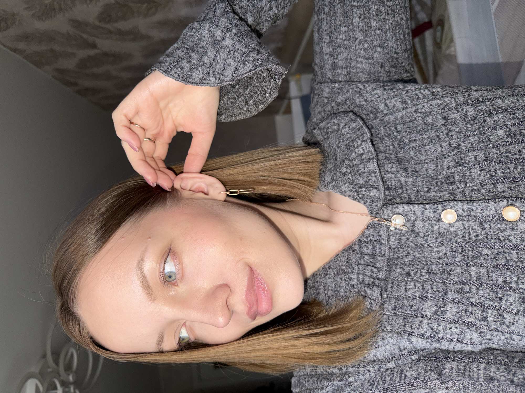 A woman with fair skin, light brown hair, and light-colored eyes is touching her ear. She is wearing a gray, textured sweater with gold buttons. Her hand has two rings. She has a gold necklace and an earring that is a gold rectangular link. The background consists of a wall, bedding, and a neutral colored wall. The location is not identifiable as Reshetnikovo, Russia based on the provided image.