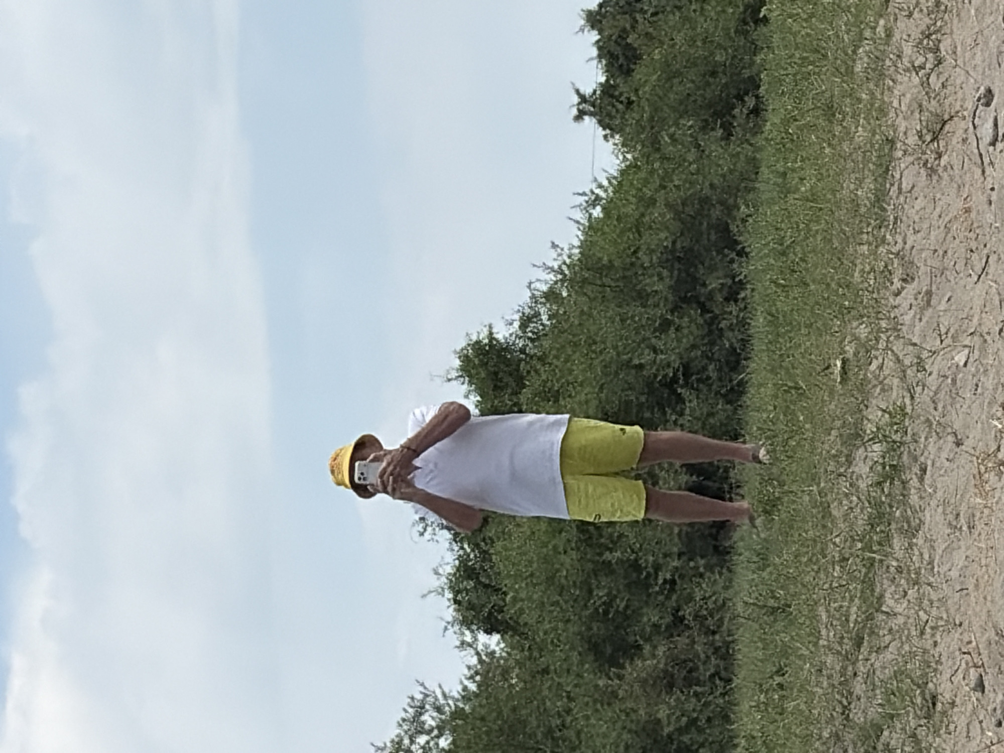 An adult male stands on a grassy area, holding a smartphone up to take a photo. He is wearing a yellow bucket hat, a white short-sleeved collared shirt, and bright yellow shorts. The man is positioned in the center of the frame, with lush green foliage behind him and a sandy foreground. The sky above is cloudy and light blue.