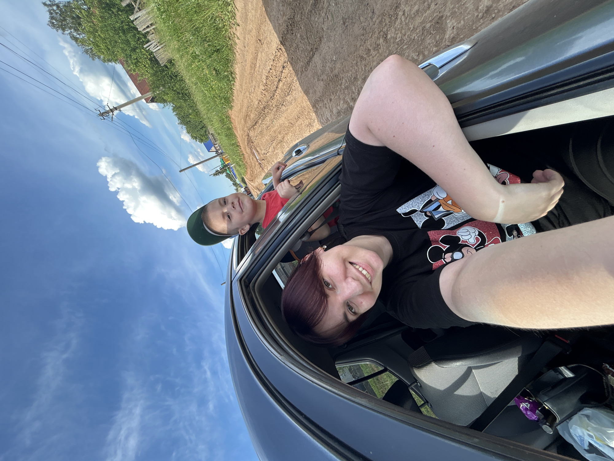 A woman and a young boy are in a car, looking out of the windows. The woman, in the foreground, is smiling and has dark auburn hair. She is wearing a black t-shirt with a graphic print of Mickey Mouse. The boy, to her left, is wearing a red t-shirt and a green baseball cap. He is looking towards the camera. They appear to be in a rural area, with a dirt road and green fields visible outside the car. The sky is blue with scattered white clouds, suggesting it is daytime and the weather is fair. The image is taken from a low angle, looking upwards. The car's interior is partially visible, showing a seatbelt and some items in the back. The scene is likely a road trip or a scenic drive in Фоки, Russia.