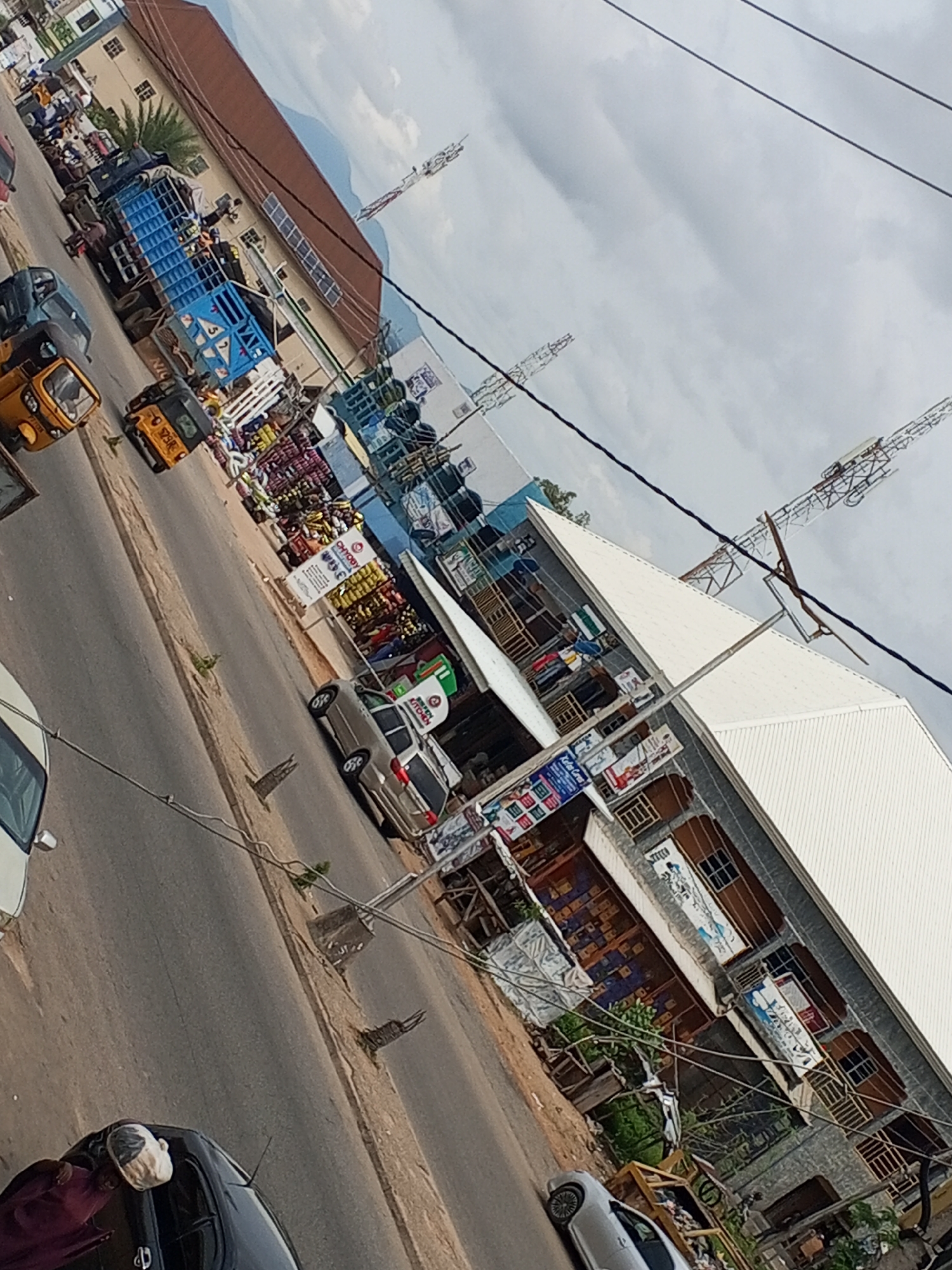 The image shows a street scene in Jalingo, Nigeria, with several vehicles and buildings. In the foreground, a black car is parked on the right side of the road. A person wearing a dark red robe and a white head covering is standing next to the car. Further down the street, a silver car is parked.
On the left side of the street, a yellow auto-rickshaw is visible, as well as a blue truck loaded with goods. Several other vehicles, including motorcycles, are parked along the roadside. Buildings line both sides of the street, some appearing to be shops with goods displayed outside. One building has a white roof and several windows. Overhead power lines crisscross the scene, and in the background, two tall radio towers are visible against a cloudy sky. The road appears to be paved, with some dirt and vegetation along the edges. The overall impression is of a busy commercial area.