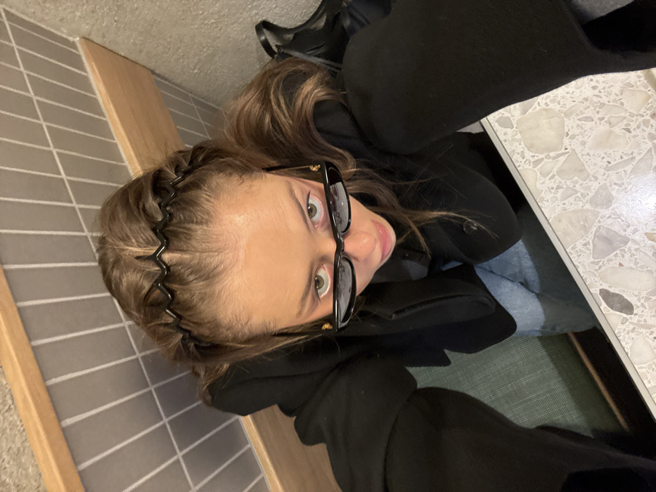 A high-angle photograph depicts a young adult female looking directly at the camera from a seated position within a commercial establishment in the city of Unknown, Unknown. She wears a black long-sleeved garment and light blue jeans. Her medium brown hair is styled with two black zigzag headbands. Dark, narrow-framed sunglasses are worn on her face, with the upper portion of her eyes visible above the frames, showing a direct gaze. Her lips are slightly parted. The background consists of a light green upholstered booth cushion, a horizontal wooden trim, and a wall paneled with grey rectangular tiles. A dark object, possibly footwear or a bag, is partially visible to the right. A speckled light-colored terrazzo countertop occupies the foreground.