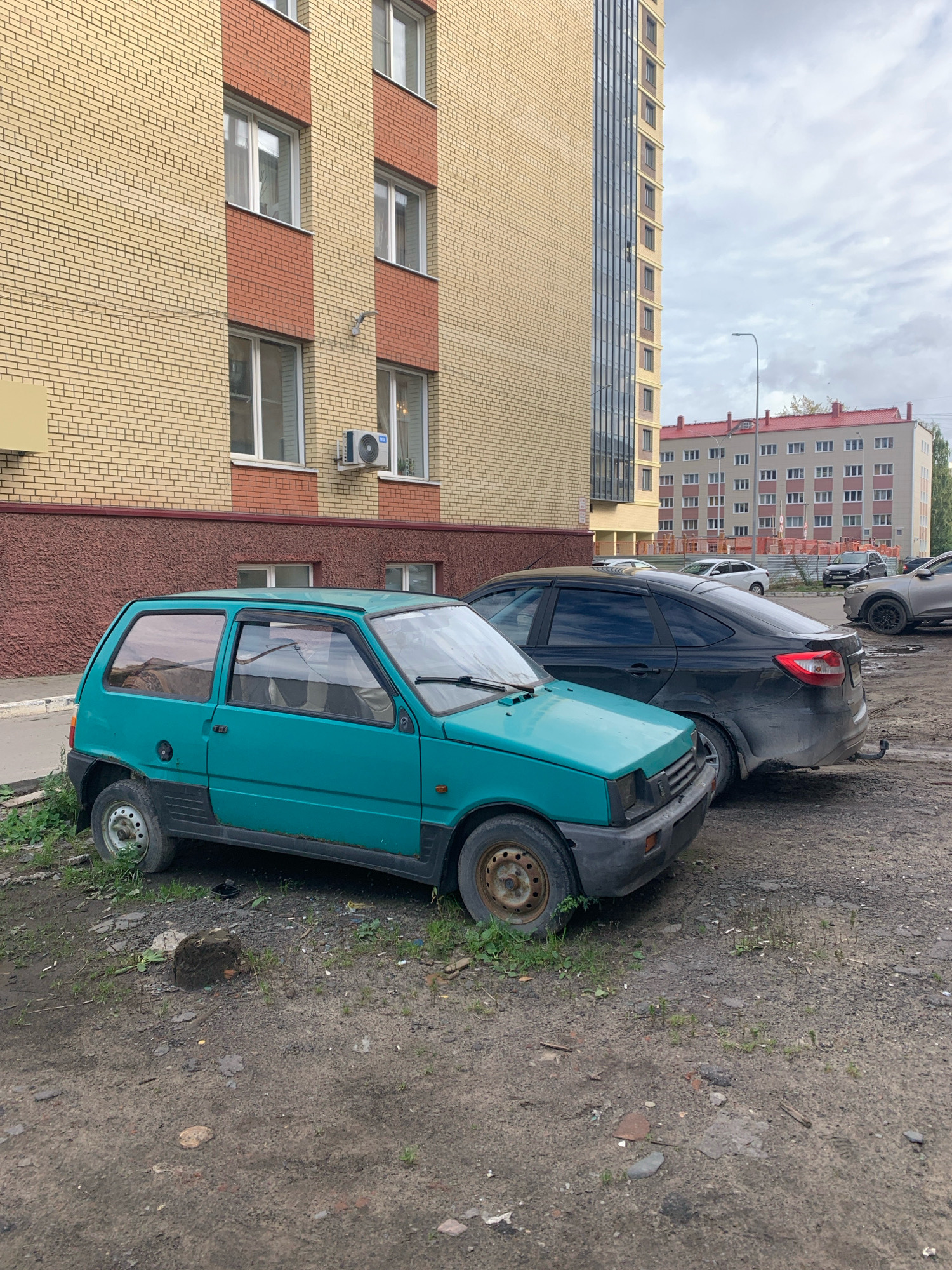 In Arkhangelsk, Russia, on a cloudy day, a turquoise hatchback car is parked on a gravel lot in front of a modern brick apartment building. The car, appearing to be a Lada Oka, is positioned closest to the camera and shows signs of wear and tear, including rust on the wheels and minor body damage. Behind it, a dark-colored sedan is parked, and further back, other vehicles and buildings are visible. The ground is covered in dirt, gravel, and some sparse vegetation. The building in the foreground has yellow brickwork with red brick accents around the windows. An air conditioning unit is mounted on the building's exterior wall.