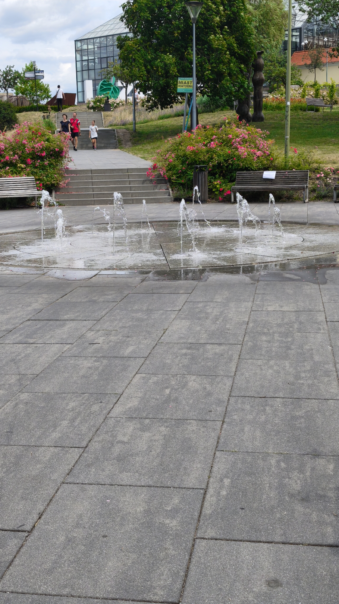 In this image, taken in an outdoor urban setting, a group of people are walking up a set of outdoor stairs. The scene features a modern glass-fronted building in the background, flanked by trees and lush green landscaping with pink flowering bushes. In the foreground, a fountain with several streams of water is active in a paved area. A wooden bench is visible to the right of the fountain. The sky is overcast.