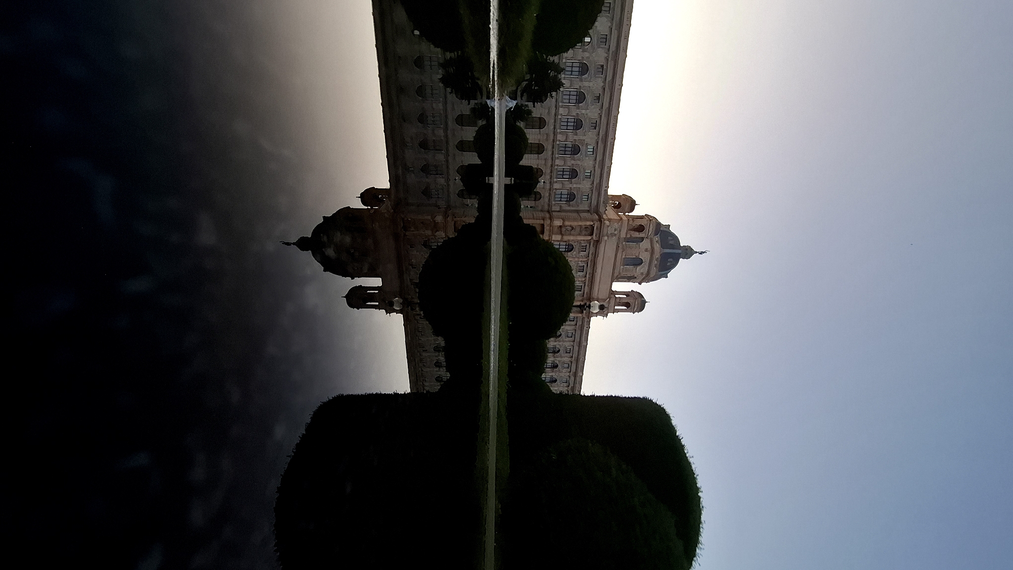 The image depicts the Kunsthistorisches Museum in Vienna, Austria, during twilight. The grand imperial building, with its distinctive dome and classical architecture, is prominently featured. In the foreground, a large body of water acts as a mirror, reflecting the museum and the dusky sky with remarkable clarity. Lush green hedges and trees are strategically placed around the building and the water's edge, adding to the scenic beauty of the setting. The sky transitions from a pale yellow at the horizon to a deep blue, indicating the early stages of night. The overall atmosphere is serene and majestic, capturing the essence of Viennese historical architecture. There are no people visible in the frame.