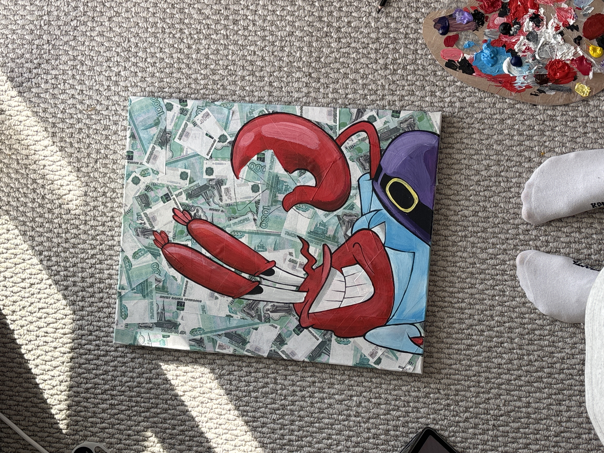 A rectangular canvas painting, oriented vertically, depicts the cartoon character Mr. Krabs from SpongeBob SquarePants, rendered in bold red, blue, purple, and yellow tones with black outlines. Mr. Krabs is shown from the chest up, with a wide, toothy grin and his large red claws extended. The background of the painting is a collage of overlapping novelty Russian 1000 Ruble banknotes, predominantly green and white, featuring "1000" denominations and Cyrillic text including "БИЛЕТ БАНКА ПРИКОЛОВ" (Ticket of the Bank of Jokes). Visible architectural features on these notes, specifically the Chapel of Paraskeva Pyatnitsa and elements resembling the Krasnoyarsk Hydroelectric Power Plant, indicate the city of Krasnoyarsk, Russia, as the symbolic location referenced.
The painting rests flat on a light grey, tightly looped pile carpet. Strong natural light illuminates the scene from the upper left, creating distinct shadows on the carpet. In the lower right corner, a wooden artist's palette is visible, covered with numerous blobs of various colored paints including red, white, black, blue, yellow, purple, and pink. Partially visible in the lower left foreground are a person's feet, clad in white ankle socks, and the hem of light grey clothing. A dark electronic device, possibly a smartphone or tablet, is partially visible on the far left edge of the carpet. A black pencil or similar tool lies near the top right edge of the palette.