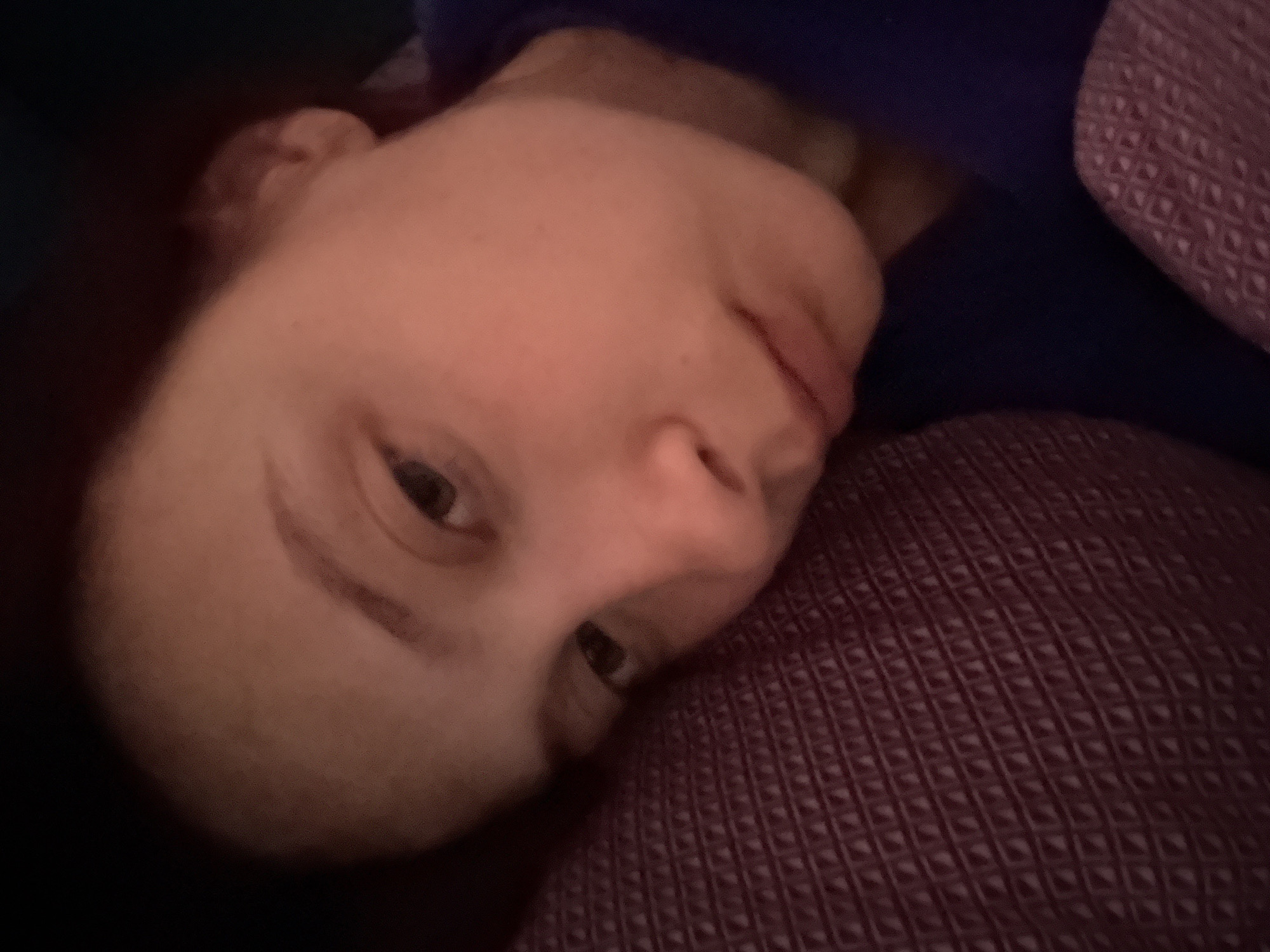 A close-up, candid shot captures a person, likely female, resting indoors in what appears to be a dimly lit setting. The image is rotated, with the individual's head tilted towards the right. Their face, with warm skin tones, occupies the central and left portions of the frame. The person's eyes are open and directed slightly upwards and to the right, conveying a neutral, perhaps thoughtful or pensive expression.
They are partially enveloped by or resting on a dark reddish-purple fabric, possibly a blanket or pillow, which features a distinct, waffle-like textured pattern. A section of dark blue fabric, possibly clothing or another covering, is visible behind their head and neck. The background is largely dark and out of focus, highlighting the person's face. The soft, low lighting suggests either evening, night, or early morning hours. No specific activity beyond resting is depicted. There is no visible text within the image. Given the provided context, the scene is situated in Прикамский, Russia.