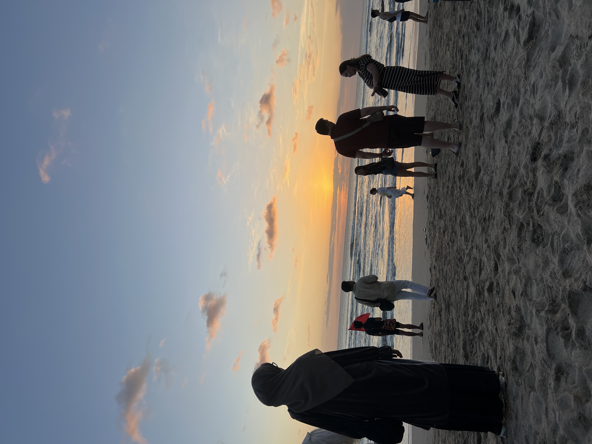 This is a full-shot, eye-level, outdoor photograph taken at dusk on the beach in Kuta, Indonesia. The sky is a gradient of blue and orange, with scattered clouds tinged with pink and orange from the setting sun. The sun is visible near the horizon, casting a warm glow over the ocean and the beach. The waves are small and gentle, lapping at the shore.
Several people are visible on the sandy beach, appearing to be tourists or locals enjoying the sunset. In the foreground, on the left, a person wearing a dark hijab and a long dark robe stands facing away from the camera, looking out at the ocean. To the right of this person, there are several other individuals, including a man in a maroon polo shirt and black shorts, and a woman in a black and white striped dress. Other figures are scattered further down the beach, some walking near the water's edge.
The sand is a light brown color and appears somewhat wet and packed near the water. The overall atmosphere of the image is peaceful and serene, capturing the beauty of a tropical sunset.