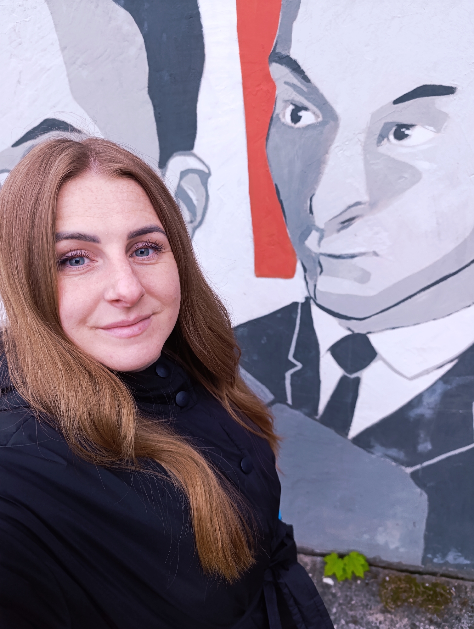 A woman poses in front of a mural on a wall. The mural depicts two men in suits, painted in shades of gray and white, with a red section in the background. The woman has fair skin, light blue eyes, and long brown hair. She is wearing a black jacket. Two small green plants grow from the base of the wall. The location is Unknown, Unknown.