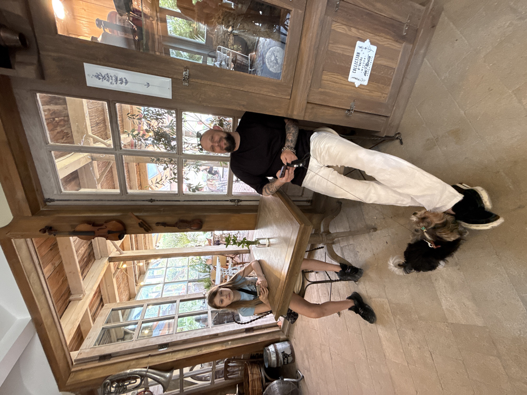 The image depicts a cozy indoor cafe scene, possibly in Semogorskiy, Russia. A young girl with long blonde hair sits at a wooden table, looking at her phone. Opposite her, a man with a shaved head and a beard is seated, also holding a phone. A small dog stands on the floor between them. The cafe has a rustic aesthetic, with exposed wooden beams, glass-paned doors, and decor that includes violins. The floor is made of large stone tiles.