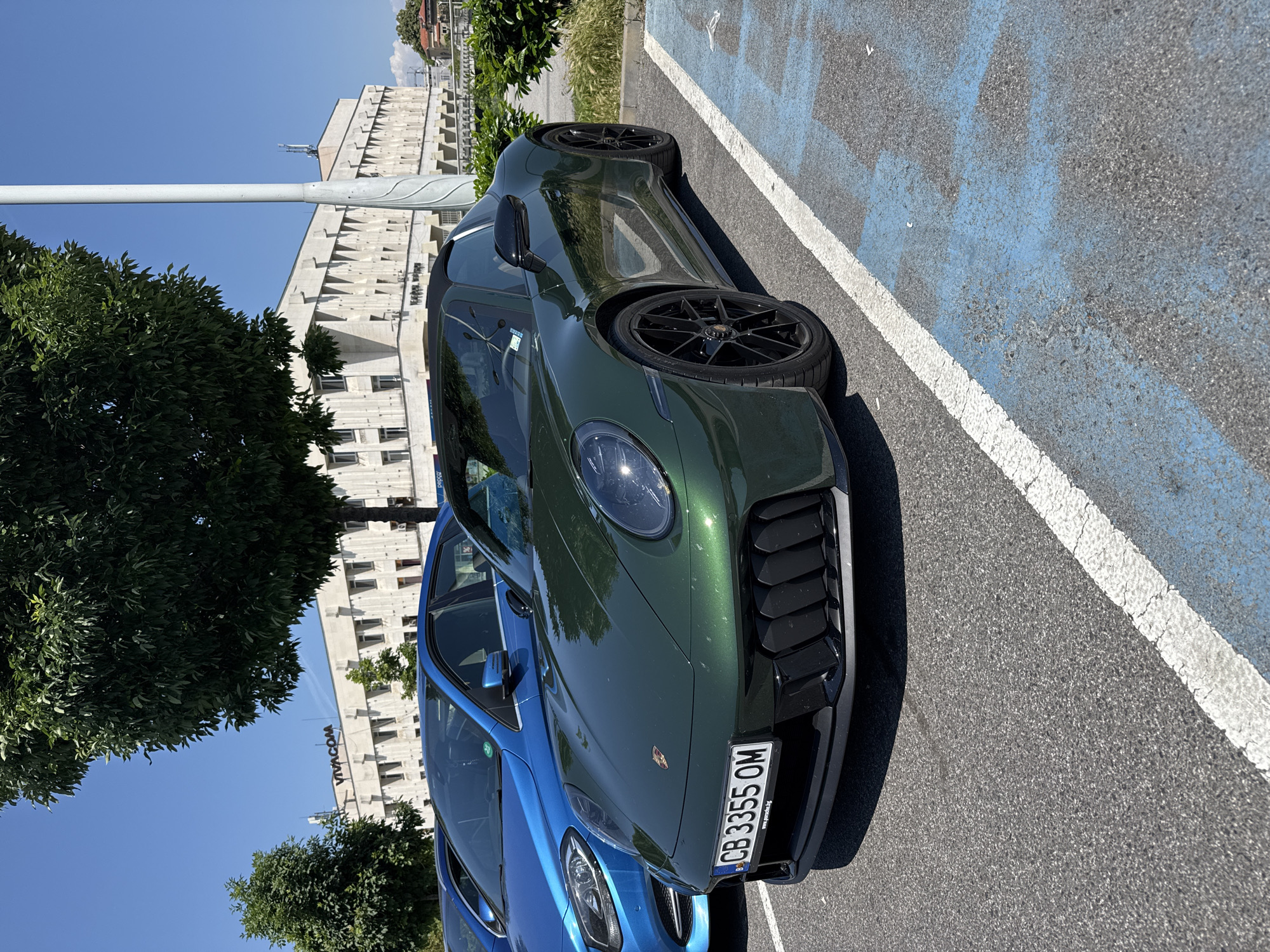 The image shows two cars parked in a designated parking spot in Plovdiv, Bulgaria. The green car, partially in focus, is a Porsche. The license plate number of the green car is CB 3355 OM. The blue car is parked to the left of the green Porsche. In the background, there's a building with "VIACOM" on the exterior, along with trees and a street light. The parking spot is marked with blue paint and a white line. The weather appears to be sunny.