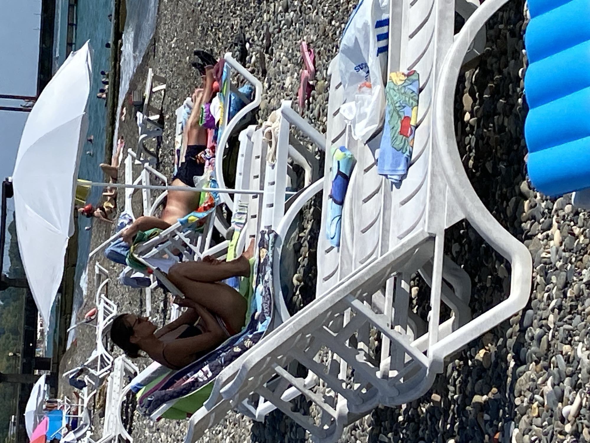 The image depicts a sunny day at a pebble beach, likely in Chabkwara, Abkhazia. In the foreground, several white plastic lounge chairs are arranged on the beach. One chair is occupied by a woman wearing a black bikini and sunglasses, reading a book. Another chair behind her is occupied by a man lying down. In the background, the turquoise sea is visible with several people swimming. A large white umbrella provides shade over one of the lounge chairs. Several beach towels and personal items are scattered on the chairs. The overall scene conveys a relaxed atmosphere of a summer vacation.