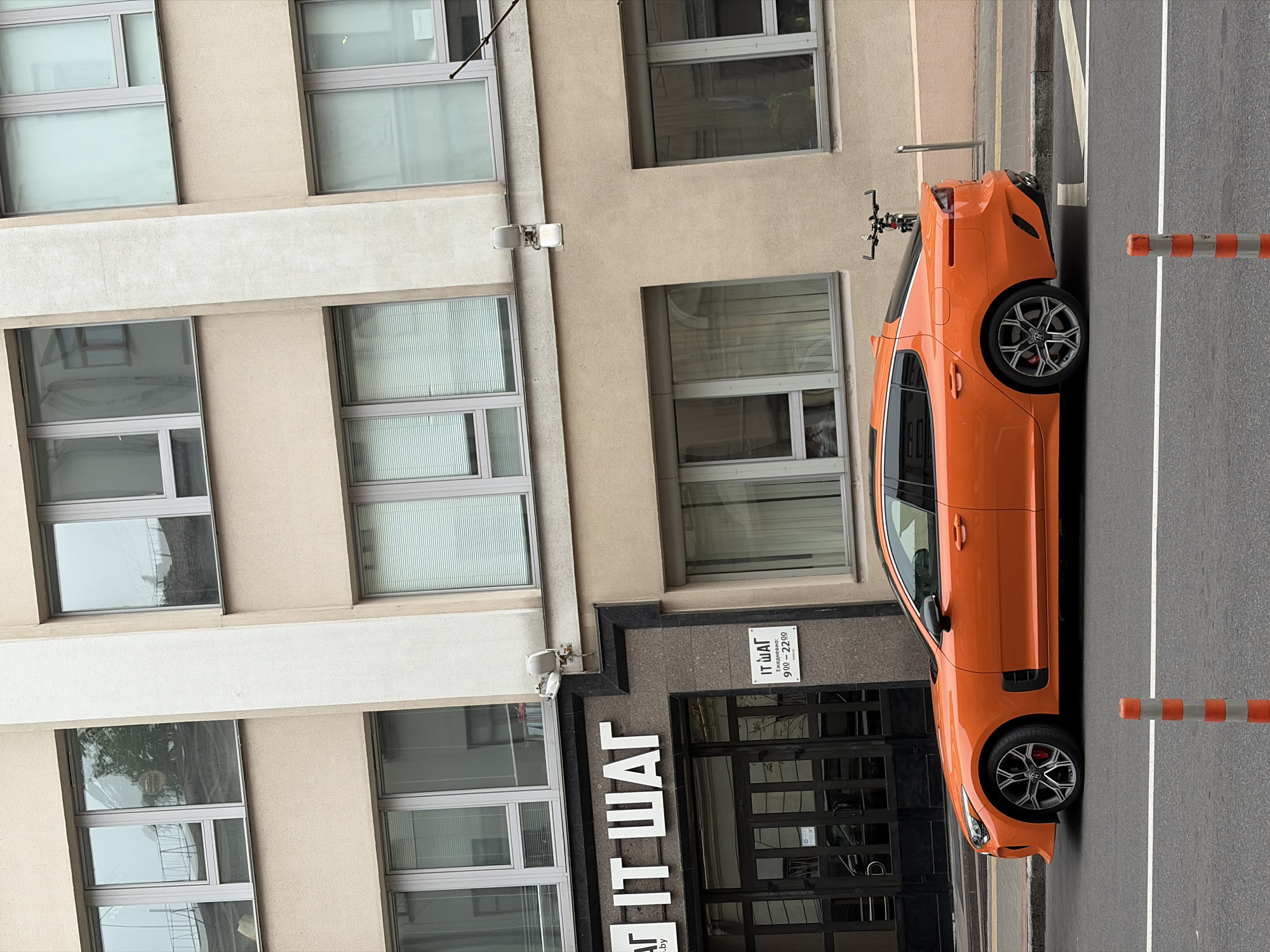 This image depicts a daytime street scene in Minsk, Belarus, specifically at Oktyabrskaya Street 5. The focal point is a multi-story building with a light beige facade and numerous grey-framed windows, some reflecting trees and structures.
On the ground floor, a dark, textured stone facade features prominent white Cyrillic letters spelling "IT ШАГ" (IT SHAG), identifying an IT academy. A smaller sign to the right of the dark-paned double entrance door reiterates "IT ШАГ" and specifies operating hours as "Ежедневно: 9⁰⁰ - 22⁰⁰" (Daily: 9:00 - 22:00). Two white wall-mounted floodlights are positioned above the entrance.
An orange Kia Stinger fastback sedan is parked parallel to the grey concrete curb on the asphalt street, facing left. The vehicle features dark multi-spoke wheels with visible red brake calipers. In the foreground, two orange and white striped traffic bollards are present on the asphalt. Behind the parked car, on the light-colored paved sidewalk, a black bicycle is leaned against the building. No individuals are visible in the scene. The overall lighting suggests an overcast day.