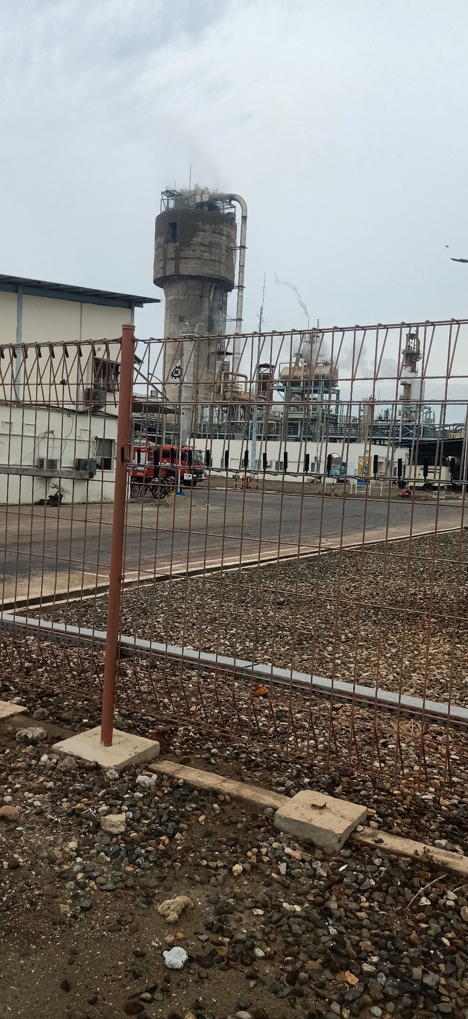 The image depicts an industrial complex under a cloudy sky, likely in Palohlada, Indonesia. A tall, cylindrical concrete structure, possibly a water tower or silo, dominates the background. Adjacent to it are various industrial buildings and pipework, with visible steam or smoke emanating from some of the structures, suggesting active operations. A red fire truck is parked in front of a building on the left side of the frame, with what appear to be motorcycles nearby. The foreground is obstructed by a metal wire fence, behind which lies a ground covered in gravel and some concrete elements. The overall scene conveys an industrial environment with a hint of preparedness, indicated by the presence of the fire truck.