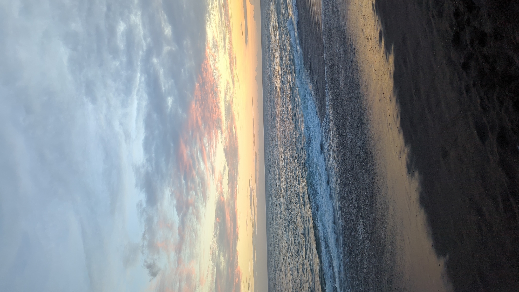 A sideways photograph shows a beach landscape in Mengwi, Indonesia. The left side of the image is filled with the sky, which features a mix of clouds. Some clouds are gray and wispy, while others are tinted with pink and orange hues from the setting or rising sun. A straight line extends across the center of the photograph, delineating the horizon where the sky meets the ocean. The ocean occupies the right side of the image. Waves are visible, crashing onto the dark sand. A strip of wet sand, reflecting the sunlight, is seen between the water and the dry sand on the far right of the frame.