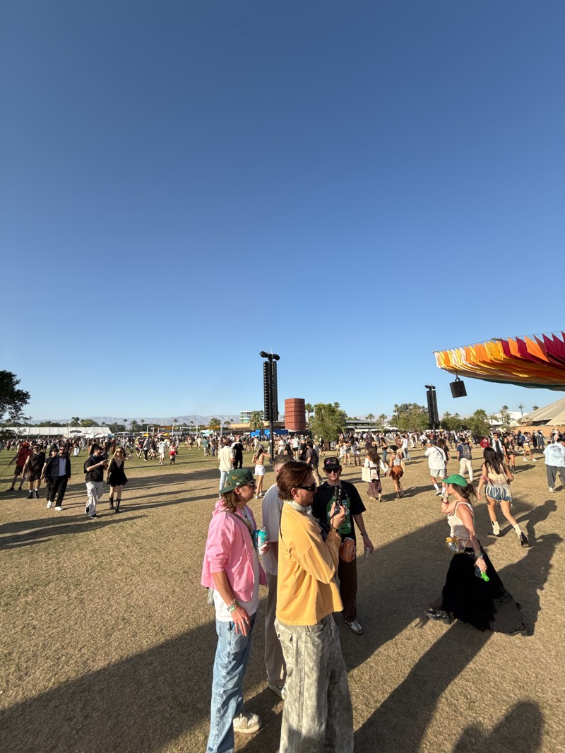 This image captures a large outdoor music festival or event taking place under a clear, bright blue sky in Indio, United States. A substantial crowd of people is scattered across a vast, dry grassy field.
In the foreground, several individuals are prominently featured, including one person in a pink hoodie and blue jeans, and another in a yellow long-sleeve shirt and patterned pants. They, along with others in the immediate vicinity, are walking, standing, and conversing. Their elongated shadows are cast on the ground, indicating it is likely mid-day to late afternoon with strong sunlight. The attendees are dressed in casual, festival-appropriate attire, suggesting warm weather.
Throughout the mid-ground and background, numerous other people are visible, moving about the field. Tall, dark speaker towers, equipped with multiple speakers, rise above the crowd, indicating the presence of a stage or performance area, although no stage is clearly visible. To the right side of the frame, a large, colorful structure made of draped, multi-hued fabric adds a distinct visual element, possibly an art installation or a decorative covering.
The setting is an open field with sparse, dry grass. In the far distance, low-lying mountains are visible on the horizon beneath the cloudless sky, characteristic of the desert landscape around Indio. Palm trees are also discernible in the background, further reinforcing the arid environment. The atmosphere appears lively and relaxed, with participants enjoying the outdoor gathering. No explicit text is visible within the image.
