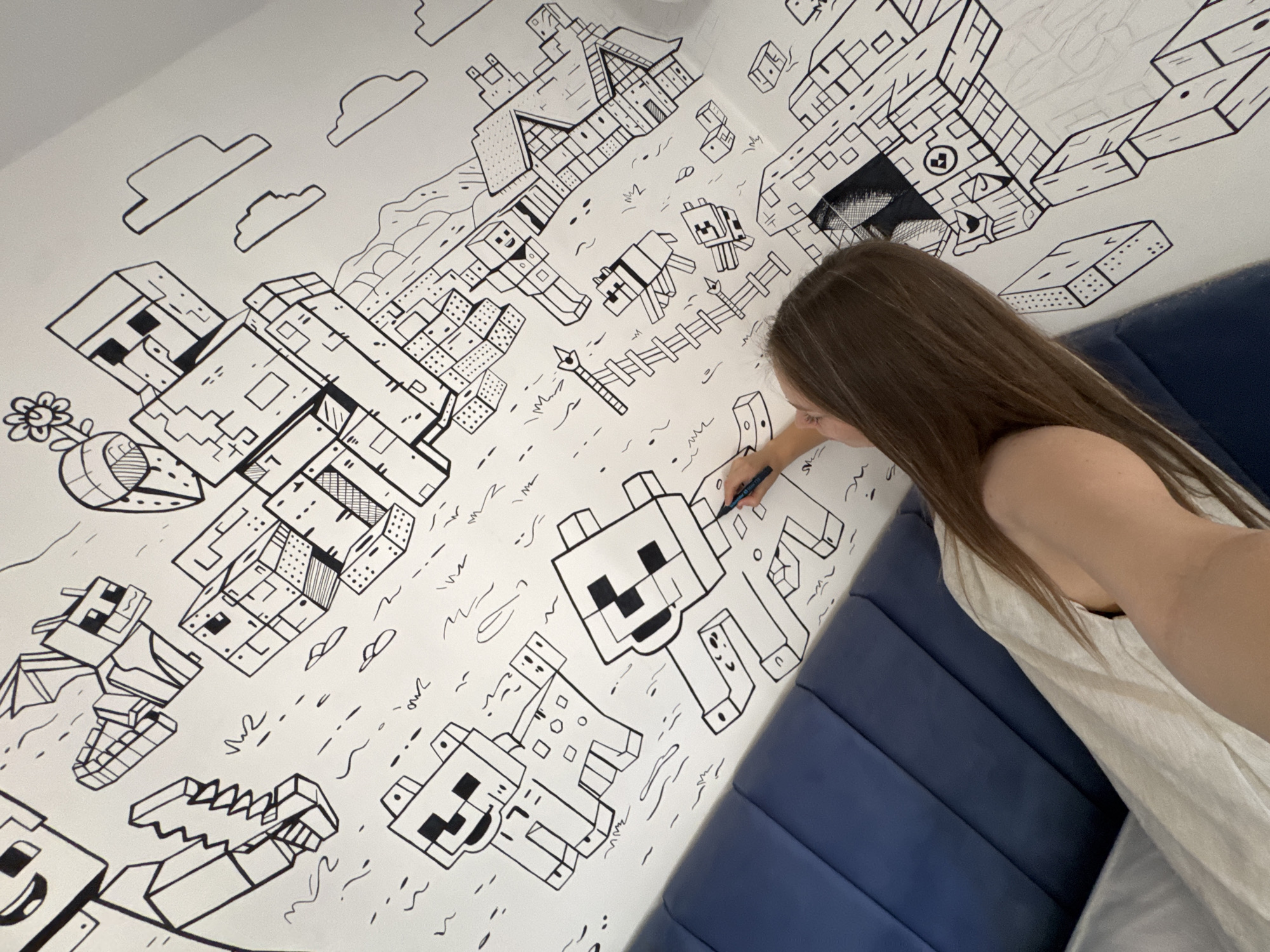 A woman is drawing a Minecraft-themed mural on a white wall. The mural includes various Minecraft characters and elements, drawn in black lines. The mural is located on a wall near a navy blue padded headboard. The woman is holding a pen or marker and is actively drawing on the mural. The location is Budva, Montenegro, but this cannot be verified from the image.