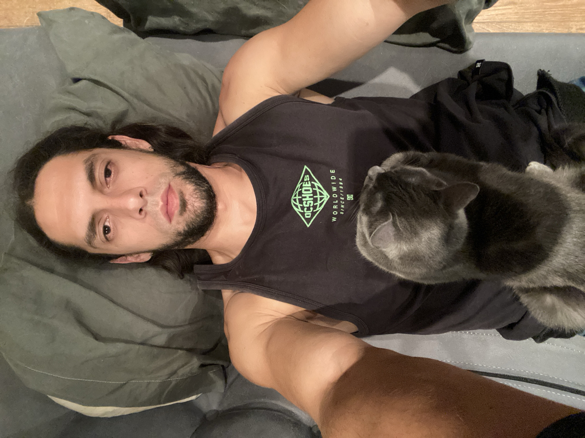 An overhead photograph captures a man lying on a dark grey surface, likely a bed or sofa, indoors in Buenos Aires, Argentina. He has long dark hair, a beard, and a mustache, and is looking directly at the camera with a neutral, calm expression. He wears a black tank top that features a prominent green diamond-shaped logo on the chest. The logo displays the text "DCSHOES WORLDWIDE SINCE 1994" along with a smaller "DC" emblem.
Resting on the man's chest is a grey-blue cat, facing away from the camera and looking towards the man's right side. The cat's fur appears soft and healthy, indicating a well-cared-for pet. The man's left arm is bent, suggesting he might be holding the camera for a selfie. The immediate background consists of dark grey pillows or bedding, while a glimpse of a wooden floor or furniture is visible in the upper right. The lighting is soft and diffused, creating a relaxed and intimate ambiance, characteristic of an evening or low-light indoor setting. The image portrays a peaceful moment of companionship between the man and his cat.
