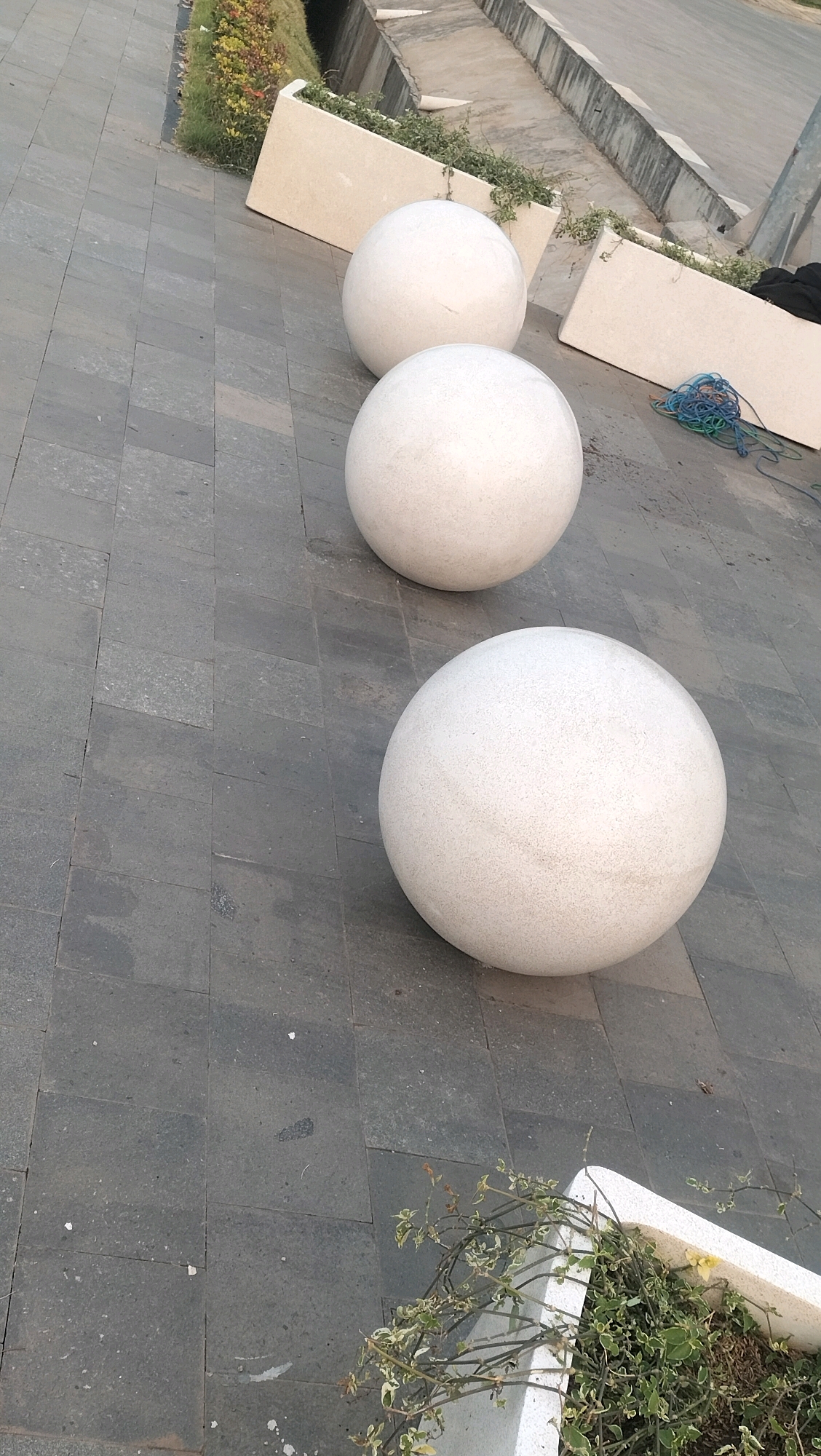 The image depicts an outdoor scene with a paved walkway constructed of large, dark gray square tiles. Arranged in a diagonal line across the walkway are three large, spherical, white decorative planters. These planters appear to be made of a textured material. Behind the planters, there are rectangular concrete planters containing some sparse greenery and soil. In the background, a concrete embankment or stairway is visible, with a hint of natural vegetation on the left side. To the right of the planters, a tangle of blue, green, and possibly brown electrical wires or cords lies on the pavement. A small amount of green foliage from a planter is visible in the bottom right corner. The lighting suggests it is daytime, possibly late afternoon, given the angle of the shadows. The image does not show any people. The scene appears to be a public or semi-public outdoor space, potentially a park, plaza, or landscaped area within a city. The location in Ketanggan, Indonesia is not visually evident from the image itself.