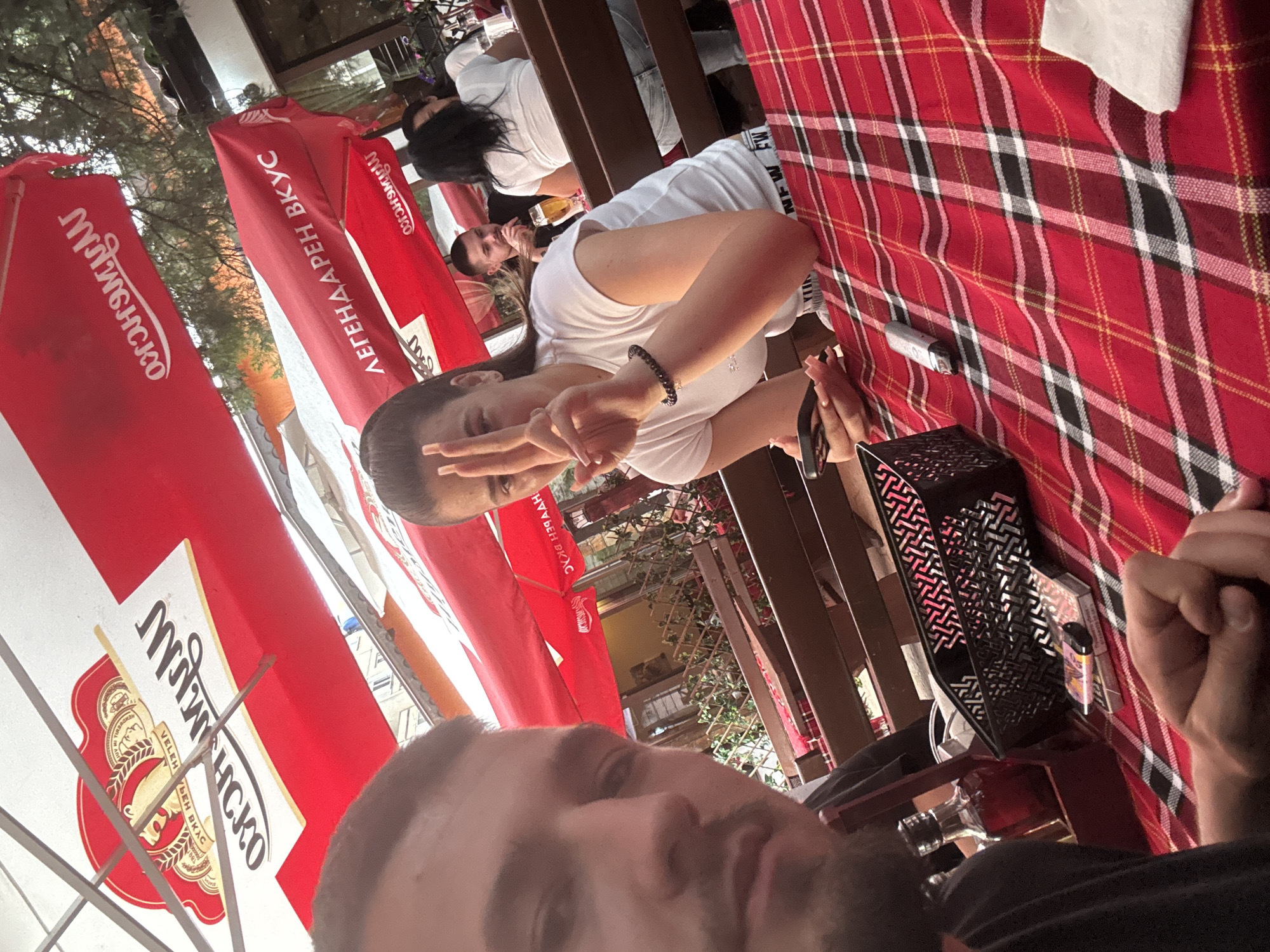 The image depicts a group of people at an outdoor cafe in Sofia, Bulgaria, under red umbrellas. In the foreground, a man is partially visible, looking towards the camera with a serious expression. A woman is seated at a table in front of him, making a peace sign with her fingers and holding a phone. She is wearing a white t-shirt and a bracelet. The table is covered with a red and white checkered tablecloth. On the table, there is a black basket, a bottle of alcohol, a pack of cigarettes, and a vape pen. In the background, other patrons are visible at tables, with one person holding a glass of beer. The overall ambiance suggests a casual dining or drinking setting. The red umbrellas bear white lettering that appears to be in Cyrillic script. One umbrella has text that translates to "Legendary Taste," and another displays a logo that is likely for a beverage brand. The environment is outdoors, with trees and foliage visible above the umbrellas.
