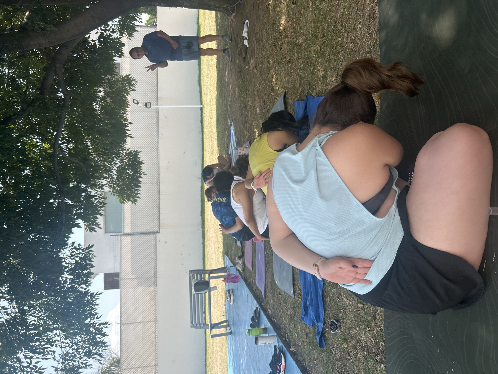 A group of people are participating in what appears to be an outdoor yoga class in Barrio de Nuevo León, Mexico. They are positioned on yoga mats laid out on the grass, with an instructor visible at the end of the row. The participants are arranged in a line, with a mix of clothing styles visible. A white building with grid-like windows stands in the background. A tree is present in the upper left portion of the image, partially shading the area. Yoga mats and personal belongings are scattered around the participants. A bench and a few water bottles are present near the yoga mats. The instructor is standing on the grass in front of the participants, gesturing towards them.