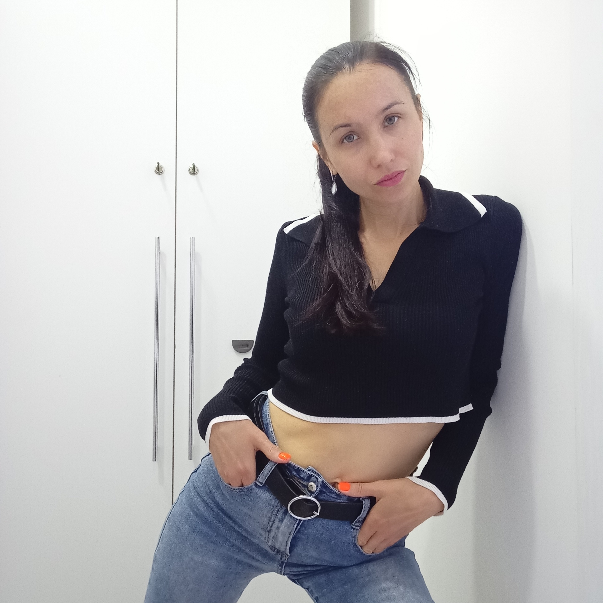A woman is leaning against a white wall next to a white wardrobe with chrome handles. She is wearing a black crop top with white trim, blue jeans with a black belt, and has dark hair pulled back. Her hands are in her jeans pockets. The location in Izhevsk, Russia is not specified in the provided image.